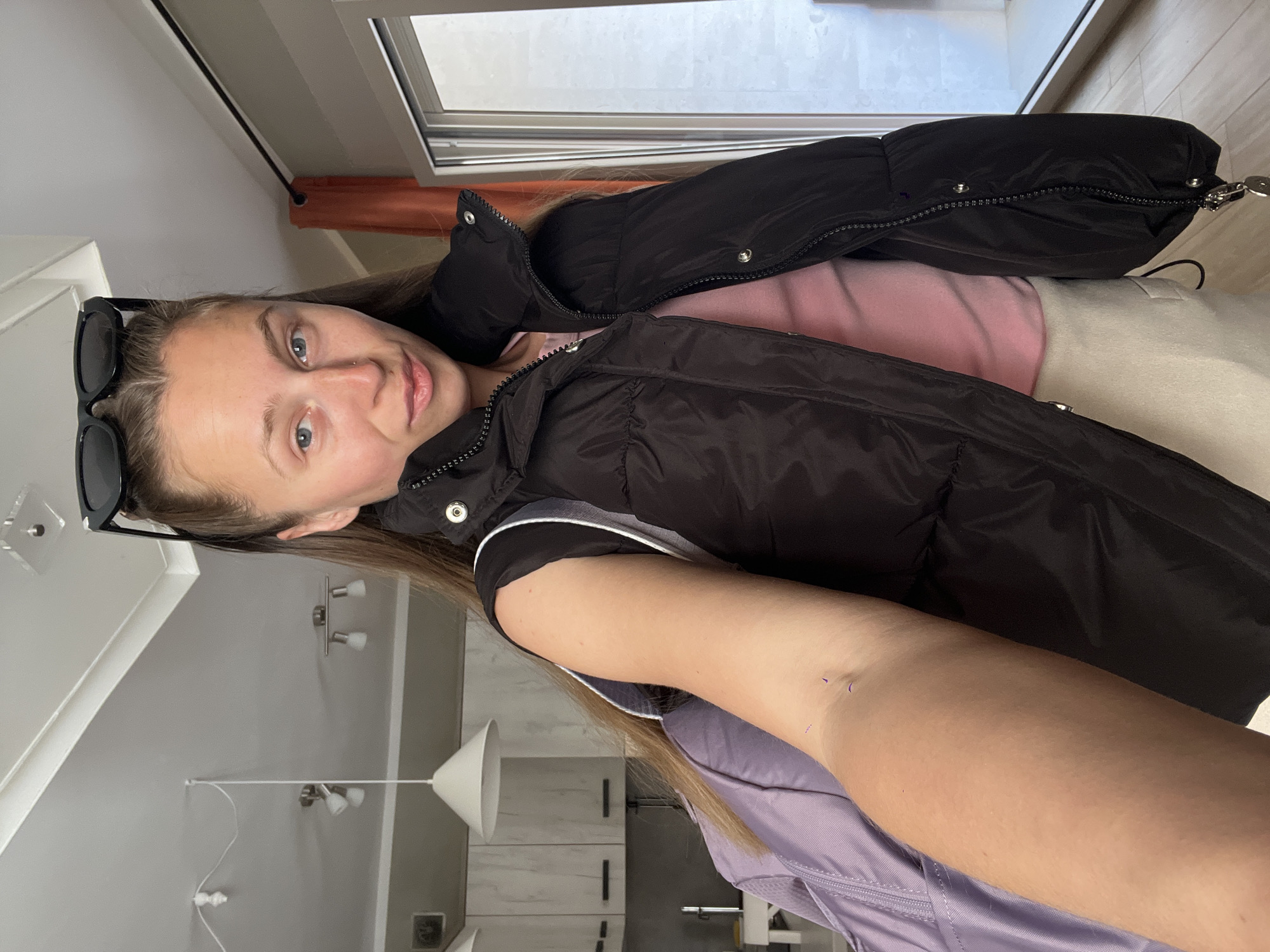 The image shows a woman in a black puffer vest over a pink shirt and light-colored bottoms. Sunglasses are propped on her head. The background reveals an interior setting with a window and light-colored walls. A white cone-shaped lamp hangs from the ceiling near a cabinet. Based on the presence of the cabinet and the lamp, it is likely a residential or indoor setting. The location in Bečići, Montenegro cannot be definitively determined from the given image as there are no identifiable landmarks or specific features that would link it to that location.