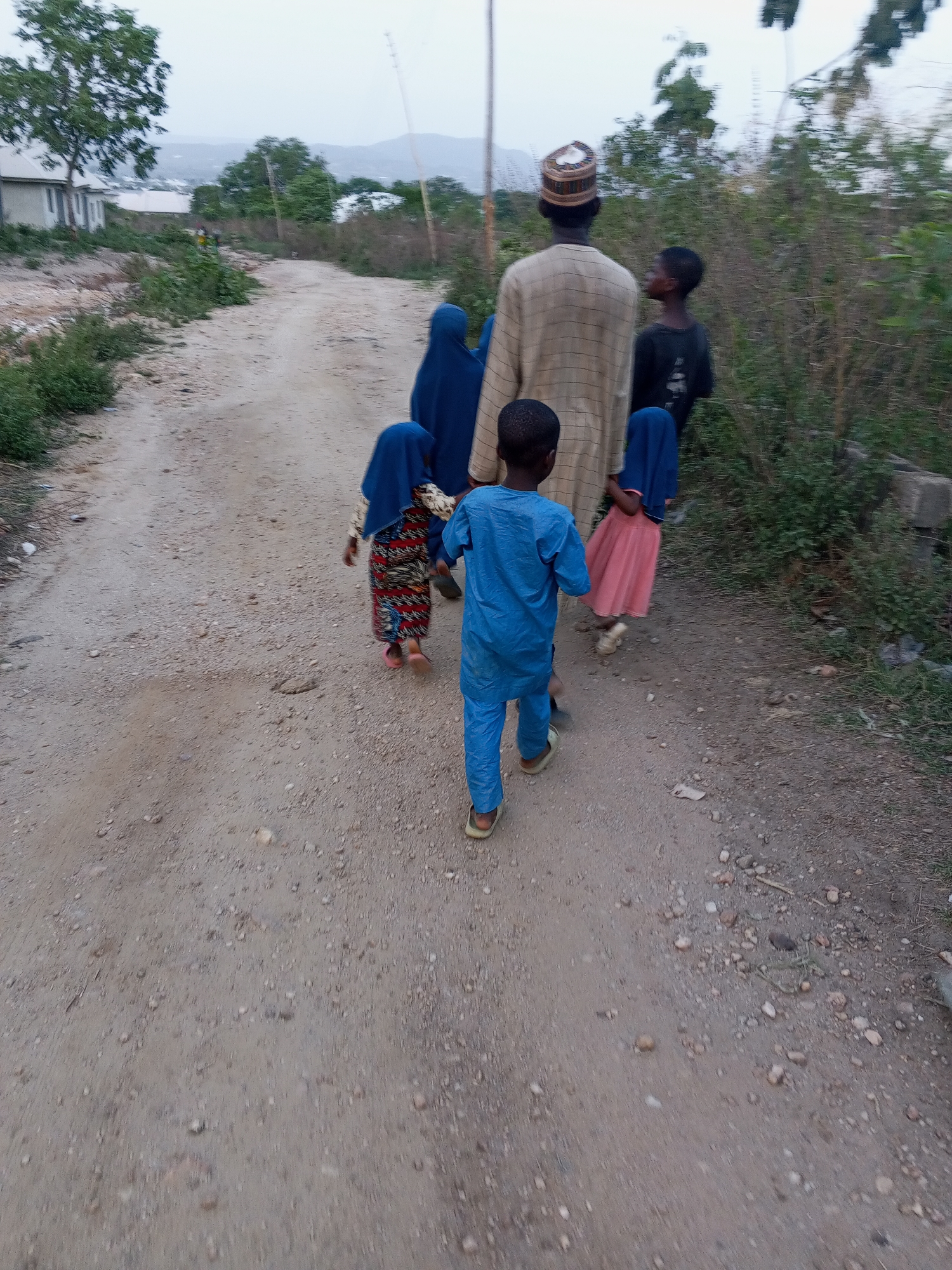 The image shows a group of people walking on a dirt road. The group consists of an adult male wearing a patterned robe and a kufi, and four children. Two of the children are wearing hijabs. The other two children are boys, one wearing a blue outfit and the other a black t-shirt. They are walking away from the camera. The background consists of vegetation and buildings. The image was taken in Zango-ndaji, Nigeria.