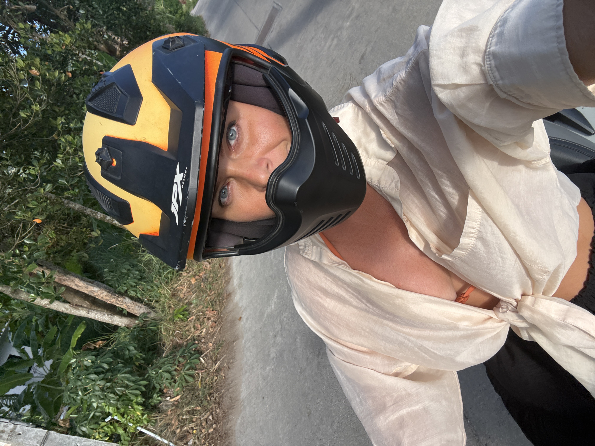 A woman with blonde hair and blue eyes is seen from the chest up, wearing a black and orange motorcycle helmet with a black face mask. She is wearing a light-colored, open-necked shirt tied at the waist, revealing an orange bikini top. Her black pants are visible at the bottom of the frame. The background shows a road and lush green foliage, suggesting a tropical environment. The lighting indicates it is daytime, with sunlight casting shadows. The image appears to be a selfie taken while riding a motorcycle. The scene is likely on a road in Ko Samui District, Thailand, given the tropical vegetation and road conditions.
