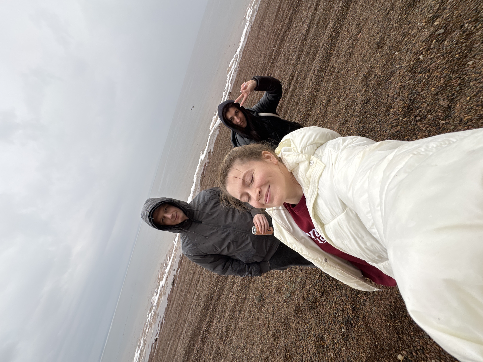 Three people are posing for a selfie on a pebble beach next to a body of water under an overcast sky. The location is likely in the city of Chaikovsky, Russia. The person closest to the camera is a fair-skinned woman with blonde hair, wearing a white puffy jacket over a red sweatshirt. To her left stands a man in a dark grey hooded jacket, holding a can. Further behind them, a woman with dark hair wearing a black hooded jacket is making a peace sign. The beach is covered in small brown pebbles. The water is visible in the background with a thin white line marking the waterline. The sky is a uniform grey.