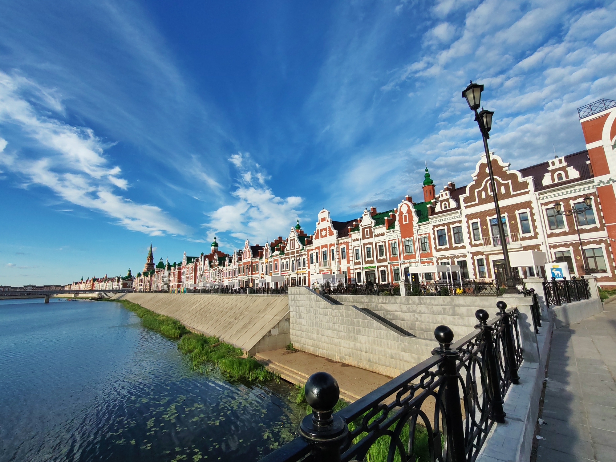 This is a wide-angle, eye-level, outdoor, daytime photograph taken in Yoshkar-Ola, Russia. The scene features a row of colorful, European-style buildings with red brick facades, white trim, and green roofs, lining the embankment of a wide, calm river. The buildings exhibit a uniform architectural style with stepped gables. A pedestrian walkway with a black wrought-iron railing runs along the river in the foreground. The river itself is a deep blue, reflecting the sky and some scattered green aquatic plants. A concrete embankment separates the river from the walkway and buildings, with some patches of green grass and reeds growing along its edge. In the distance, a bridge spans the river. The sky is a vibrant blue, filled with wispy white clouds. A lamppost with two lights stands on the walkway, near the buildings. The overall impression is one of a well-maintained, picturesque waterfront area.