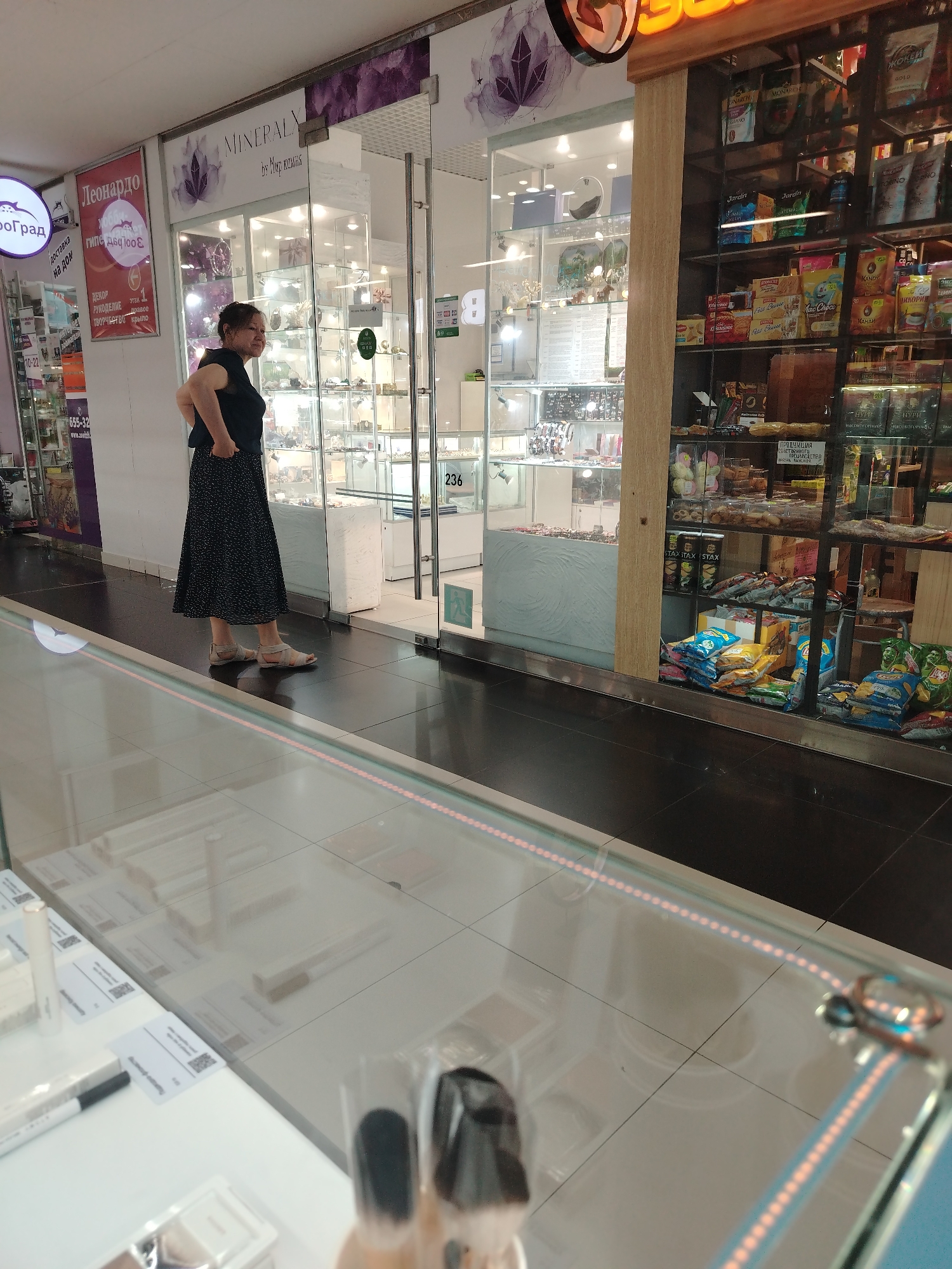 A middle-aged woman wearing a dark top and a black and white polka dot skirt stands in front of a storefront in what appears to be a shopping mall. The store on the left, labeled "MINERAL," features glass displays with various items inside. Adjacent to it on the right is another store selling packaged foods and snacks.
In front of the woman, there is a glass display case. The image also includes various signs and labels in Russian, including "Леонардо" and "Доставка на дом."
Based on the signs, the location appears to be in the city of Izhevsk, Russia.