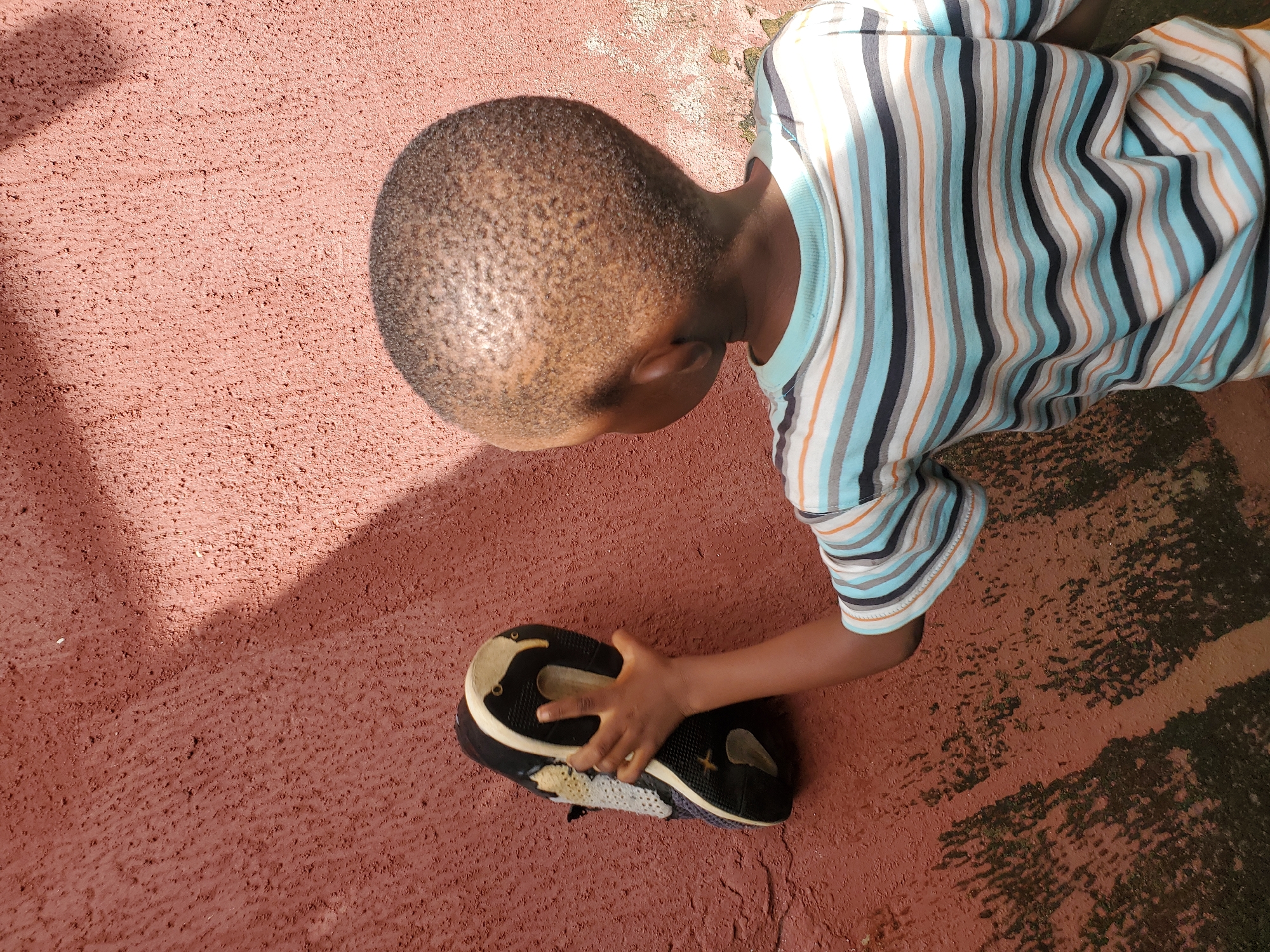 A young Black boy, viewed from behind, is pressing a dark-soled sneaker with a light-colored sole against a rough, reddish-brown wall. He wears a horizontally striped t-shirt with alternating bands of blue, white, orange, and black. The wall appears textured and weathered, with some dark, possibly damp, patches and streaks on its lower half, suggesting an outdoor urban environment. Sunlight casts a distinct shadow on the wall, indicating it is daytime. The scene is likely taking place in Jos, Nigeria, a city known for its hilly terrain and often vibrant, textured architecture. The action appears to be playful or exploratory, with the child interacting with the wall and the shoe as props.