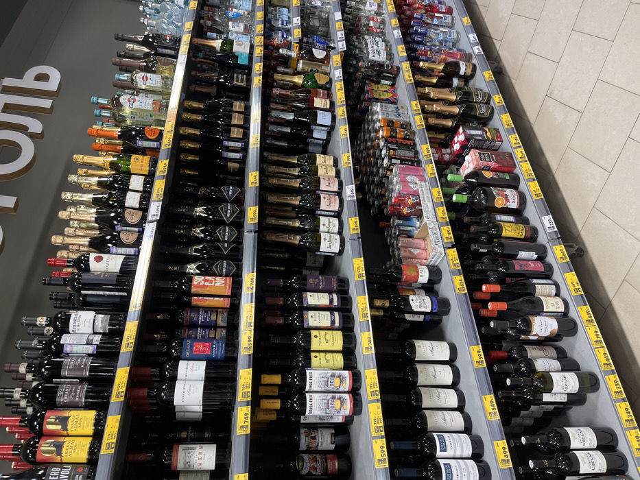 The image displays a brightly lit alcohol aisle within a retail establishment in Ширяево, Russia. Multiple dark gray metal shelving units are fully stocked with a wide variety of alcoholic beverages arranged on at least six tiers. The shelves are predominantly filled with bottles of red wine, sparkling wine, champagne, and various spirits, including vodka, gin, rum, and liqueurs. Brands such as 'TIERRA ALMA', 'CHIANTE', 'MARTINI', and 'SOLANO' are identifiable on bottle labels. Yellow price tags with black lettering, displaying prices in Russian Rubles (e.g., 799.00, 1499.00, 599.00), are affixed to the front edge of each shelf, positioned beneath their respective products. Lower shelves also feature canned beverages, including various beers, ciders, and mixed drinks, displayed in cardboard trays and individually. A large, white, uppercase Cyrillic sign, partially visible and reading "ОЛЬ" (likely part of "АЛКОГОЛЬ"), is mounted on a dark wall above the uppermost shelf. The floor is composed of light-colored, square tiled sections. No individuals or discernible actions are present in the scene, which captures a static view of merchandise within a retail environment.
