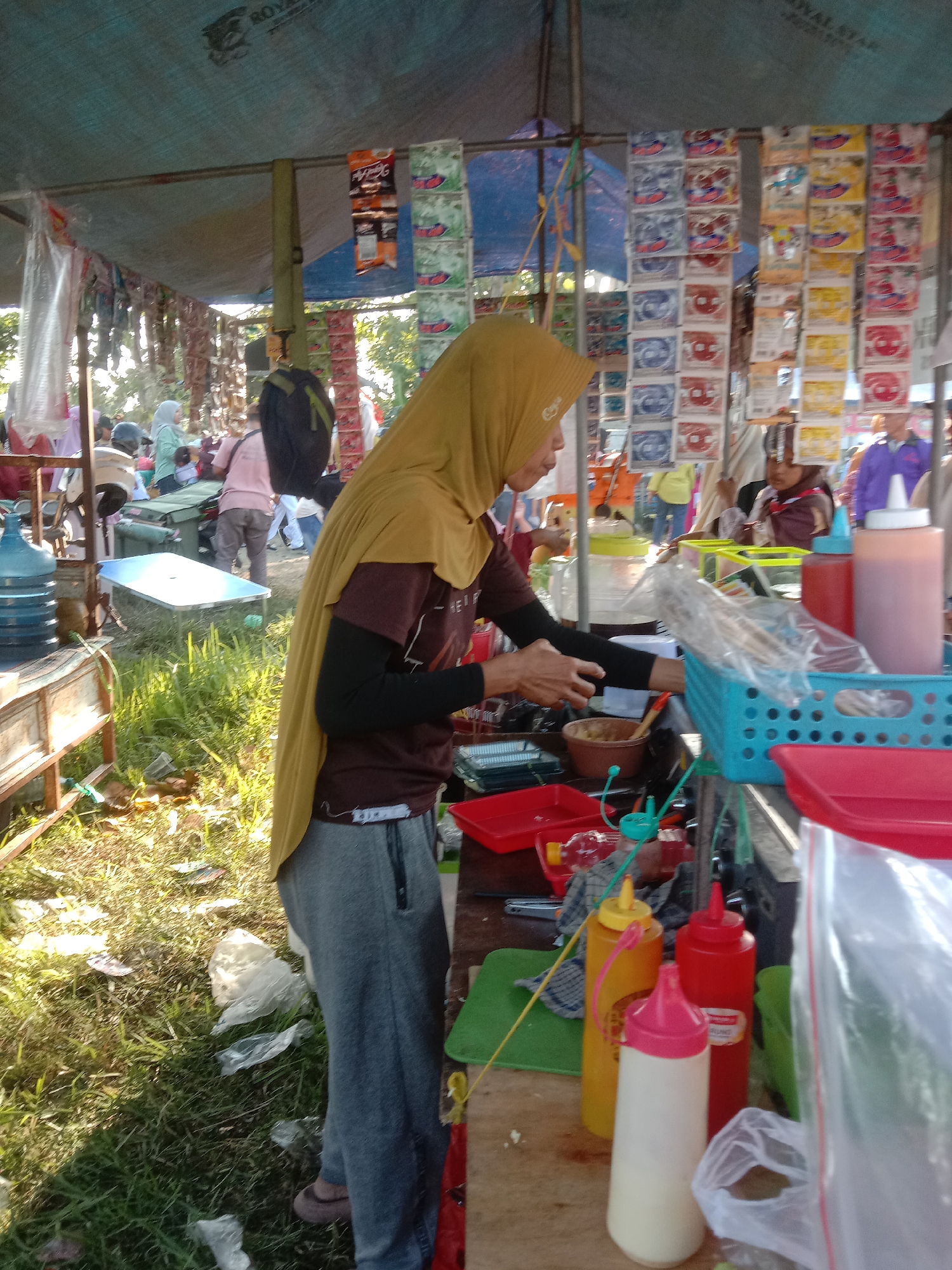 This image captures a lively street food stall, likely in the city of Banda Aceh, Indonesia, given the cultural context of the scene. The stall is set up under a blue tarp, offering shade from the sun. The vendor, a woman wearing a yellow hijab and a dark maroon shirt with black sleeves, is actively preparing food, her hands engaged in the process. She is surrounded by various food preparation items, including red trays, a blue plastic basket filled with skewers, and an assortment of condiment bottles, one red, one yellow, and a white one with a pink cap. Behind her, the stall displays a vibrant collection of colorful product packaging, possibly snacks or candies, suggesting a bustling marketplace atmosphere. Other individuals are visible in the background, some appearing to be customers or fellow vendors, contributing to the dynamic scene. The ground is covered in grass, with some scattered plastic debris, indicating an outdoor, possibly informal, setting. The overall impression is one of a busy commercial area in a tropical climate.