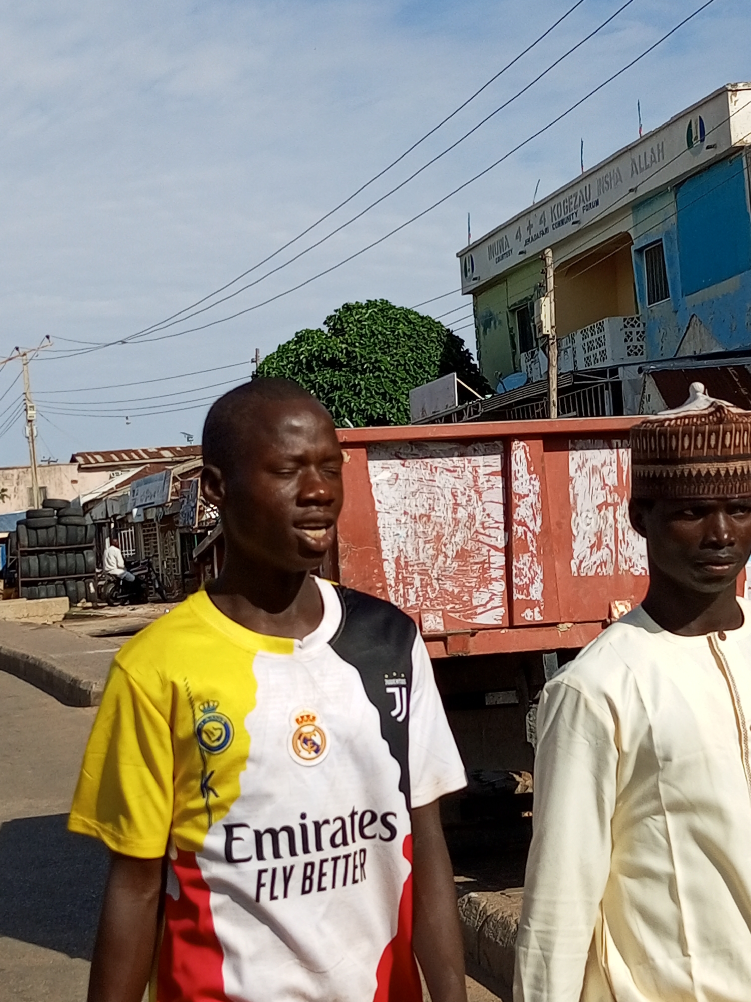 In the city of Arawa, Nigeria, two young men are captured in a street-level photograph. The man on the left, closer to the camera, is wearing a distinctive t-shirt with a yellow, white, and black color-block design, featuring logos from both Real Madrid and Juventus. He has his eyes closed and his mouth slightly open, suggesting he might be speaking or mid-action. To his right, another young man is visible, wearing a light-colored traditional tunic and a patterned cap. He is looking towards the viewer.
Behind them, a large, bright red truck is parked, its side covered in peeling white posters. Further in the background, a two-story building displays a sign with text, and other smaller structures, possibly market stalls or workshops, are visible. A pile of stacked tires and a motorcycle with a rider are also present in the scene, indicating a busy urban environment. Overhead, power lines crisscross the sky under a clear, light-blue sky. The overall scene depicts a moment in the daily life of Arawa, Nigeria.