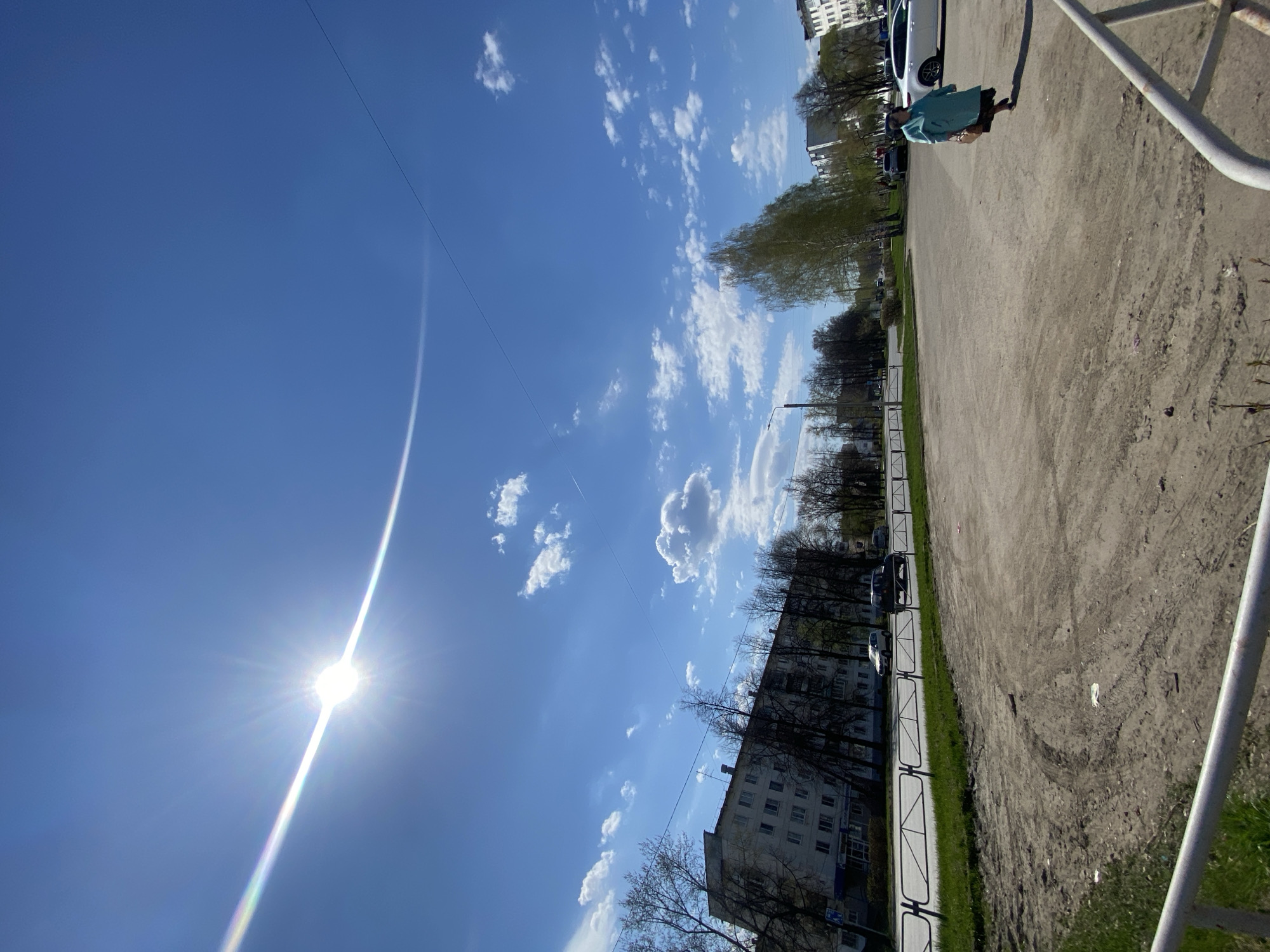 The image depicts a sunny day in Chaikovsky, Russia. The sky is blue with scattered white clouds and the sun is shining brightly. A power line stretches across the upper part of the sky. A multi-story building is seen along the left side, and there are trees with bare branches. On the right, there's a dirt and concrete surface with a white metal railing. A person wearing a turquoise top and a dark skirt is partially visible on the right. Cars are parked along the street.