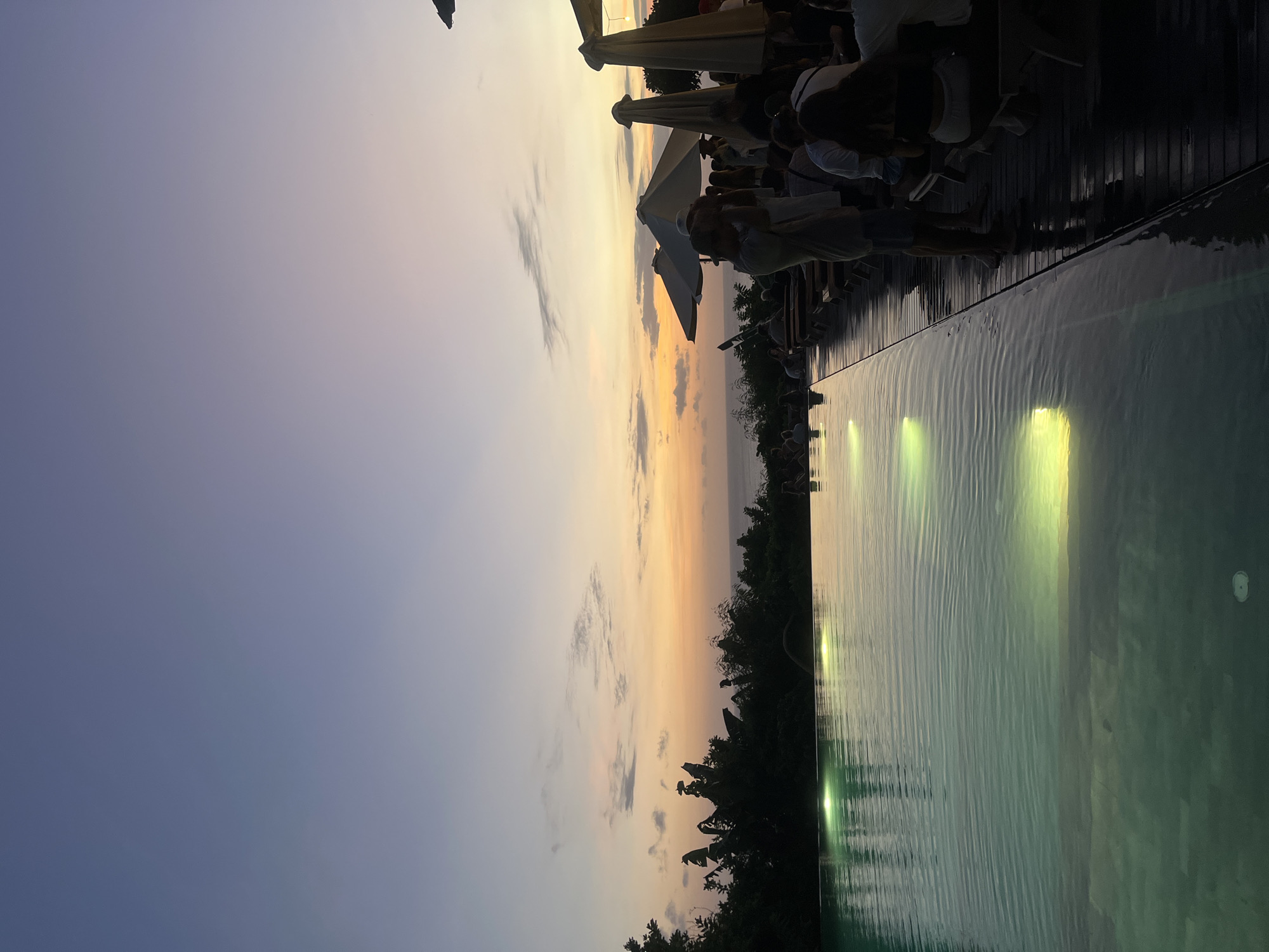 The image, taken in Kuta Selatan, Indonesia, shows a view at dusk looking towards the horizon over an infinity pool. The pool is lit from below with lights that reflect on the water surface. People are seated at tables under umbrellas on a deck beside the pool. Silhouette-like trees, possibly palm trees, are visible along the horizon line. The sky is a gradient of colors, with blue at the top, transitioning to orange and yellow near the horizon. Clouds are scattered throughout the sky.