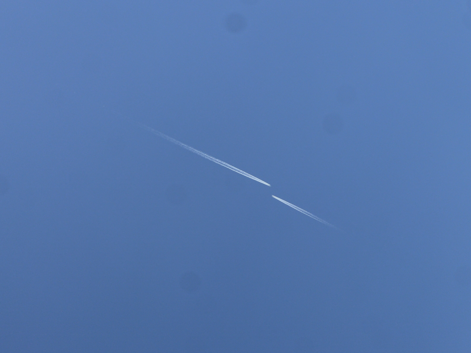 The image shows a clear blue sky over Bar, Montenegro. Two distinct contrails, likely from aircraft, diagonally cross the frame. The contrails appear as thin, white lines fading at their ends, indicating distance. The scene is devoid of any other objects or people.