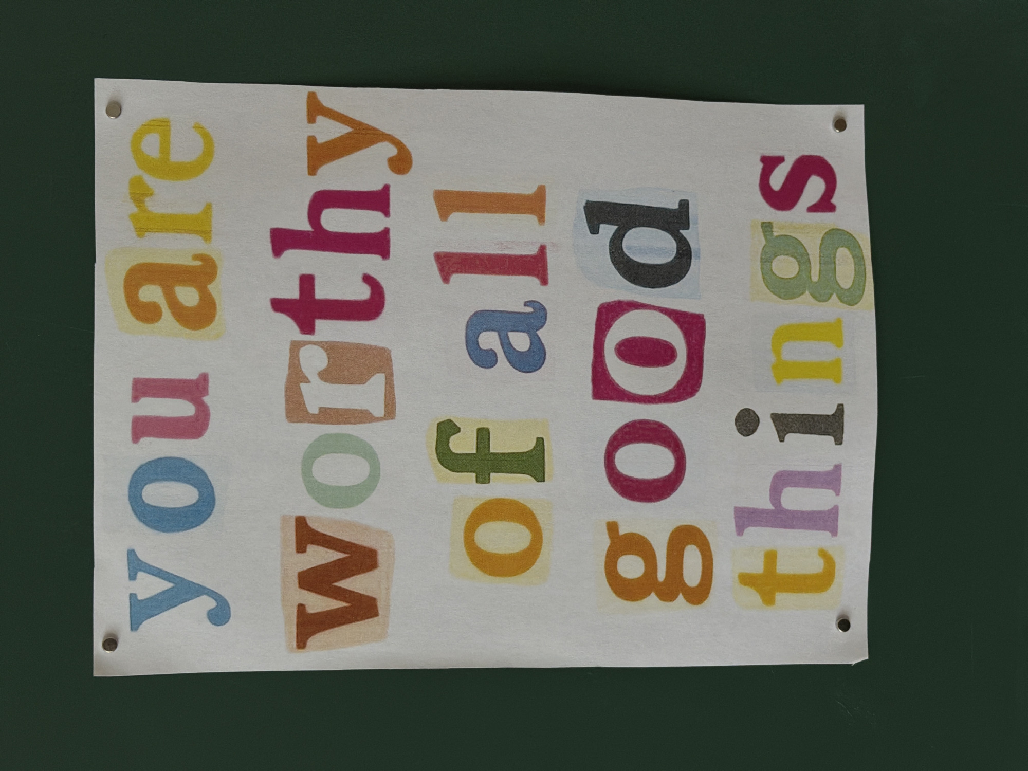 A motivational poster hangs crookedly on a green background. The text on the poster, created with a collage of colorful, cutout letters, reads "You are worthy of all good things." The poster is attached at the top corners with small, silver-colored fasteners.