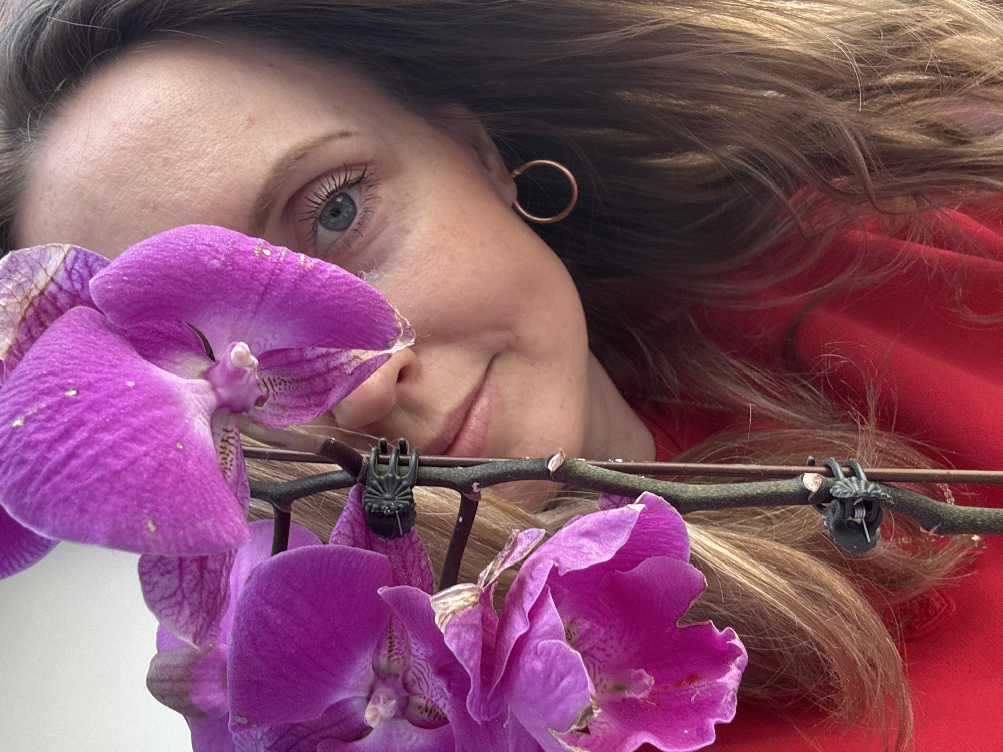 A close-up photograph features a woman with fair skin, light brown hair, and blue eyes, partially obscured by purple orchid flowers. She wears a red garment and a simple, copper-colored hoop earring. The orchid stem is visible, with green clips attached. The background is blurry, making it difficult to discern the environment. There is no definitive information in the image to confirm the location is Chaikovsky, Russia.