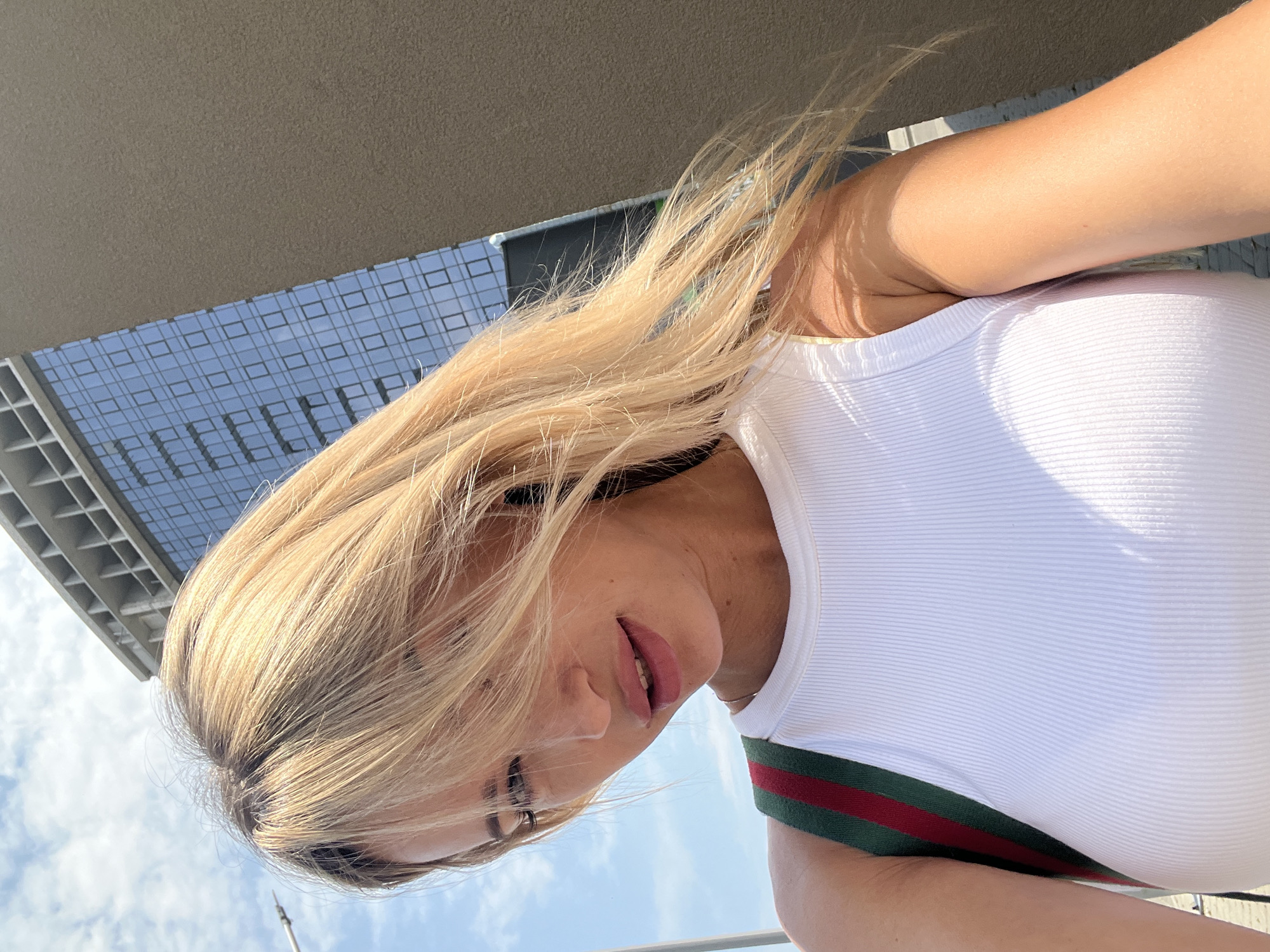 A woman with blonde hair and light skin is taking a selfie outdoors. She is wearing a white ribbed tank top and a bag strap with red and green stripes. The background shows a building with blue glass windows and a cloudy sky. The image appears to be taken in the city of Fontanka, Ukraine.