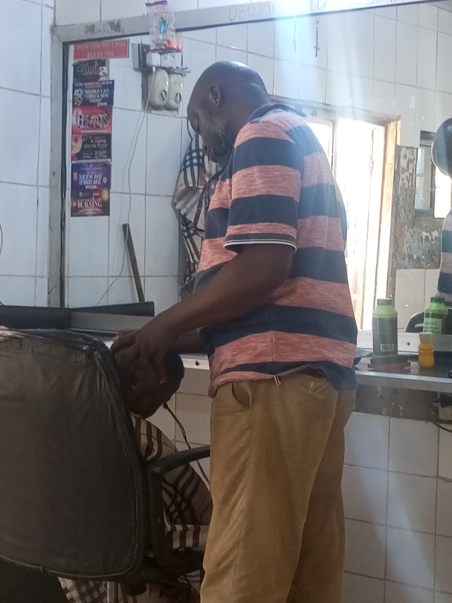 A man with a shaved head and short grey beard, wearing a striped shirt and tan trousers, is working on a customer in what appears to be a barbershop or salon. The customer is seated in a black chair. The room has white tiled walls with posters and signs. There are grooming products visible on a counter. The environment is consistent with the city of Yelwa, Nigeria.