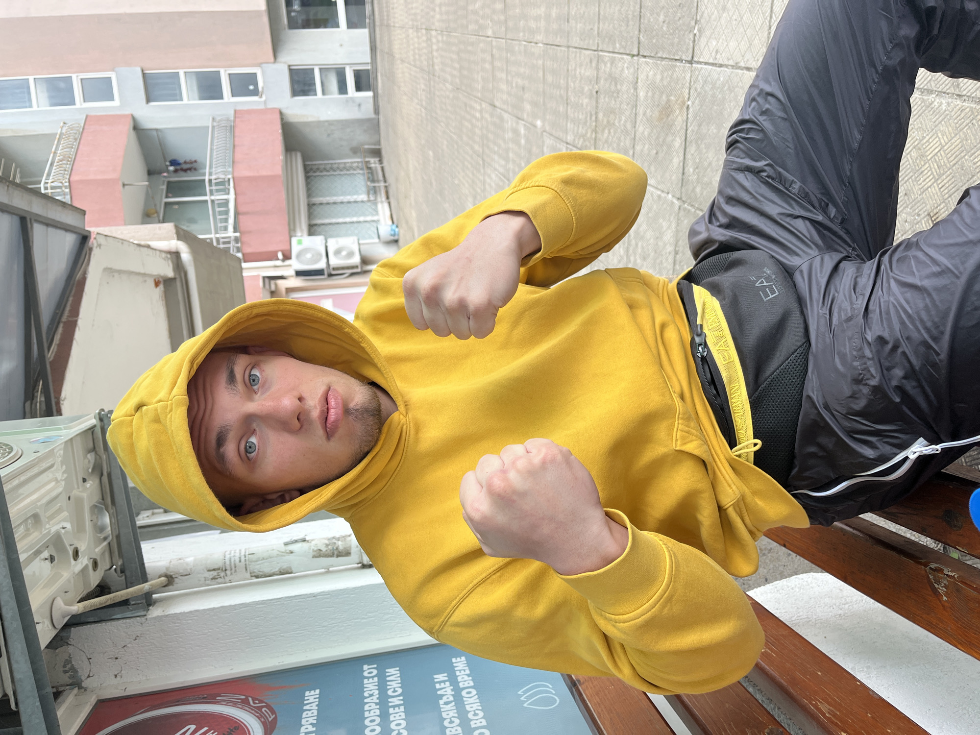 A young man sits outdoors in Burgas, Bulgaria, appearing to pose or joke for the camera. He wears a yellow hooded sweatshirt and black track pants. His fists are clenched, as if to box. He is positioned on a wooden bench. Behind him is a light-colored concrete exterior and a building with windows. There's also signage with writing in Bulgarian.