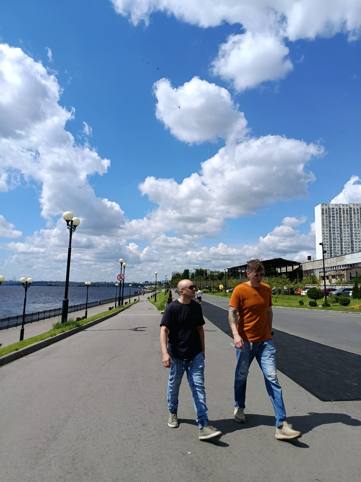 Two men walk along a paved promenade adjacent to a wide river under a bright blue sky filled with fluffy white clouds. The man on the left is bald and wears a black t-shirt and ripped blue jeans. The man on the right wears a bright orange t-shirt with visible tattoos on his left arm and ripped blue jeans. Both men are wearing casual shoes and appear to be walking in the same direction. The promenade is lined with lampposts and a black metal fence separates it from the river. In the background, across the river, there are buildings and industrial structures. To the right of the promenade is a road with sparse traffic, bordered by trees and green spaces, and a tall, modern white building. The scene suggests a warm, sunny day in an urban setting. The location is identified as Saratov, Russia.