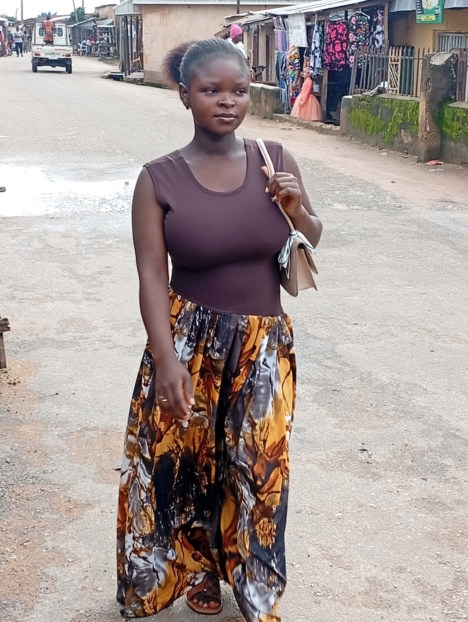 A young woman with dark skin and her hair in a bun is walking down a dusty, unpaved street in a market area in Jos, Nigeria. She is wearing a dark brown tank top and a long, flowing skirt with a bold, abstract print in shades of brown, yellow, black, and white. She is carrying a light-colored handbag over her left shoulder. Her left hand is resting on her hip, and she is wearing a ring on her left ring finger. Her feet are bare, with one foot partially visible wearing a brown sandal.
The background shows a typical Nigerian market scene. Buildings with corrugated metal roofs line the street, with stalls displaying various goods, including clothing. People are visible in the distance, including individuals walking and a white truck with a person standing on its back. The street is worn and dusty, indicating a well-trafficked area. The overall atmosphere suggests a bustling, everyday scene in a Nigerian city.
