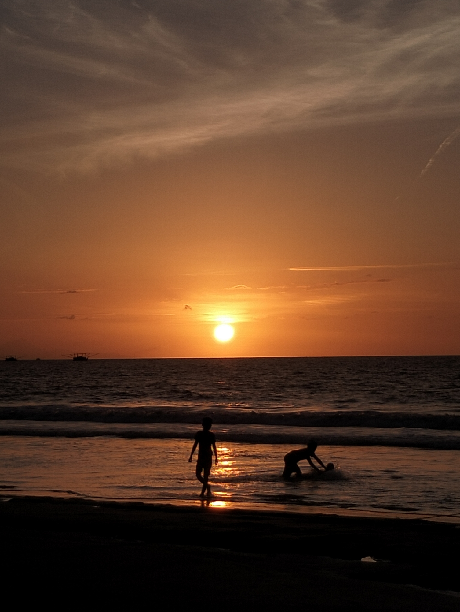The image depicts a silhouetted scene at dusk on a beach in Uleekareung, Indonesia. Two children are present in the shallow water. One child stands facing the ocean, while the other kneels, appearing to interact with a ball. The sun is low on the horizon, casting a warm orange glow across the sky and water, with clouds streaking across the upper portion of the frame. The ocean has moderate waves. In the distance, two fishing boats are visible on the horizon. The foreground consists of dark sand.