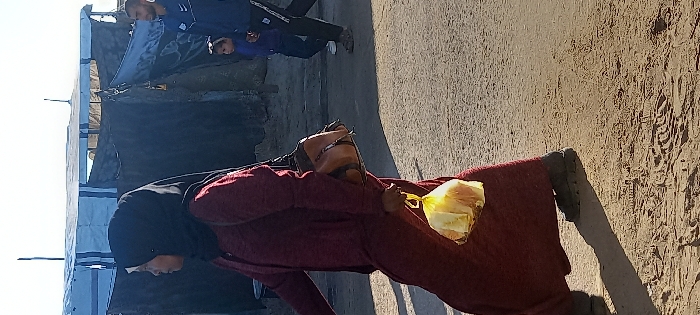 A woman wearing a black hijab and a long maroon dress walks across an unpaved dirt and gravel path in Khan Yunis, Palestinian Territory. She carries a tan shoulder bag on her right side and holds a transparent yellow plastic bag containing light-colored items, possibly bread, in her right hand. Her shadow extends onto the ground in front of her. In the mid-ground to the right, a man in a dark blue athletic jacket with white detailing and dark pants walks towards the left, accompanied by a small child in a dark blue top. Both the man and child are partially visible. The background features temporary structures, including a white and blue striped tarp tent and darker fabric shelters, under a clear sky. Tire tracks and footprints are visible in the foreground's dirt ground.