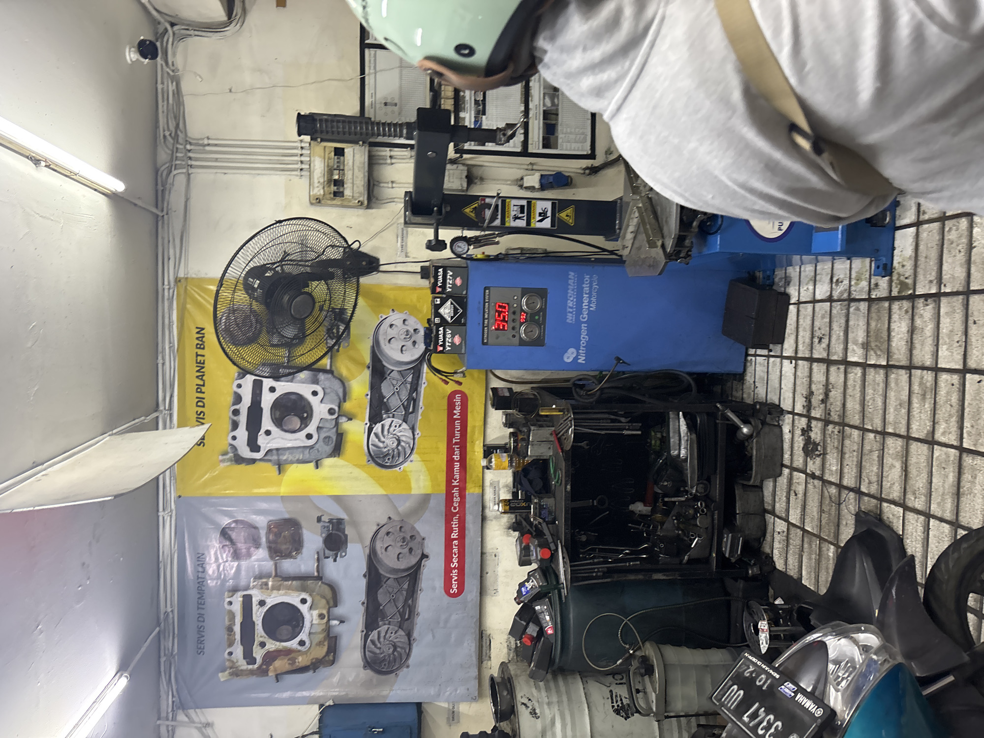 The image depicts the interior of a motorcycle repair shop in North Jakarta, Indonesia. The scene is cluttered with various tools, equipment, and spare parts. In the foreground, a portion of a turquoise and black Yamaha motorcycle is visible, with a license plate reading "3347 UU". To the right, a person wearing a light gray hoodie and a mint green helmet is partially visible from behind, seemingly observing the machinery.
Dominating the center of the image is a blue "NITROMAN Nitrogen Generator Motorcycle" machine with a digital display showing "35.0" and "992". Adjacent to this machine is a black industrial fan and a rack filled with assorted tools. In the background, two large posters are mounted on the wall; one on the left is a gray poster with images of motorcycle engine parts and text in Indonesian, while the poster on the right is yellow with similar engine part illustrations and text. Several electrical conduits and a fuse box are visible on the wall. The floor is tiled with a grid pattern, showing signs of wear and dirt. The overall environment suggests a functional, working workshop.