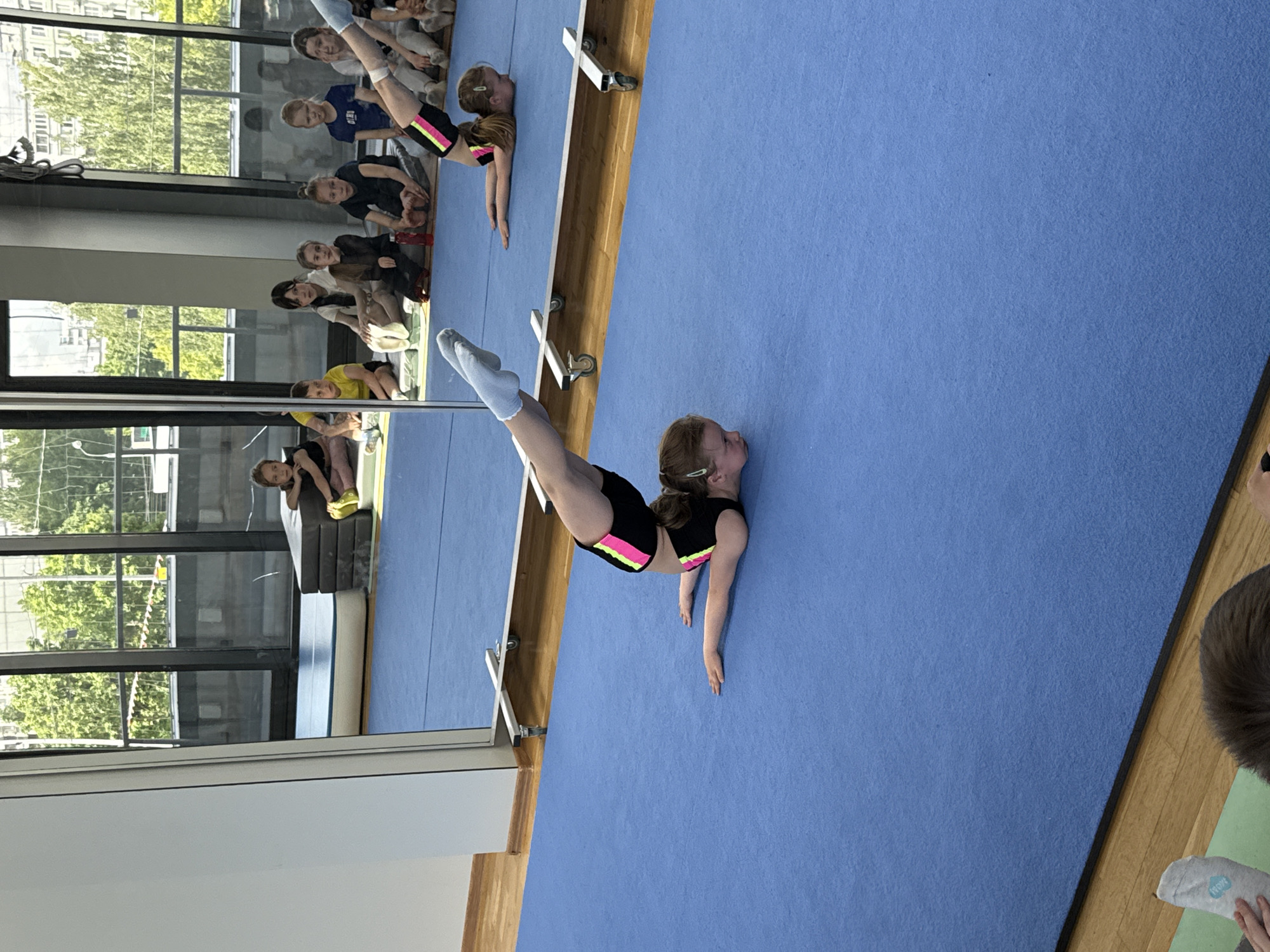 The image depicts an indoor gymnastics or dance studio, likely in Khimki, Russia, bathed in natural light, suggesting it is daytime. The primary focus is a young girl with light brown hair, dressed in a black athletic top and shorts with neon pink and yellow accents, performing a gymnastic pose on a large blue mat. She is lying on her stomach, arching her back with her head lifted, and her legs are extended vertically upwards. Her hands are on the mat, supporting her pose.
To her left, a large mirror reflects the room. Visible in the reflection are several other young girls, likely participants or observers, sitting or crouching along a wooden base beneath the mirror. One reflected girl is seen wearing a blue t-shirt with the white Russian word "СЛОН" (SLON), meaning "elephant," printed on it. Another reflected girl is also in the process of performing a similar arched back and leg extension pose.
The background features expansive windows that look out onto an urban environment, revealing green trees and buildings, indicating an outdoor view from the facility. In the bottom right corner, a foot wearing a light green sock or on a light green mat is partially visible, with faint, partially obscured text that may read "WOU" or similar. The scene conveys a focused training or practice session for young gymnasts.
