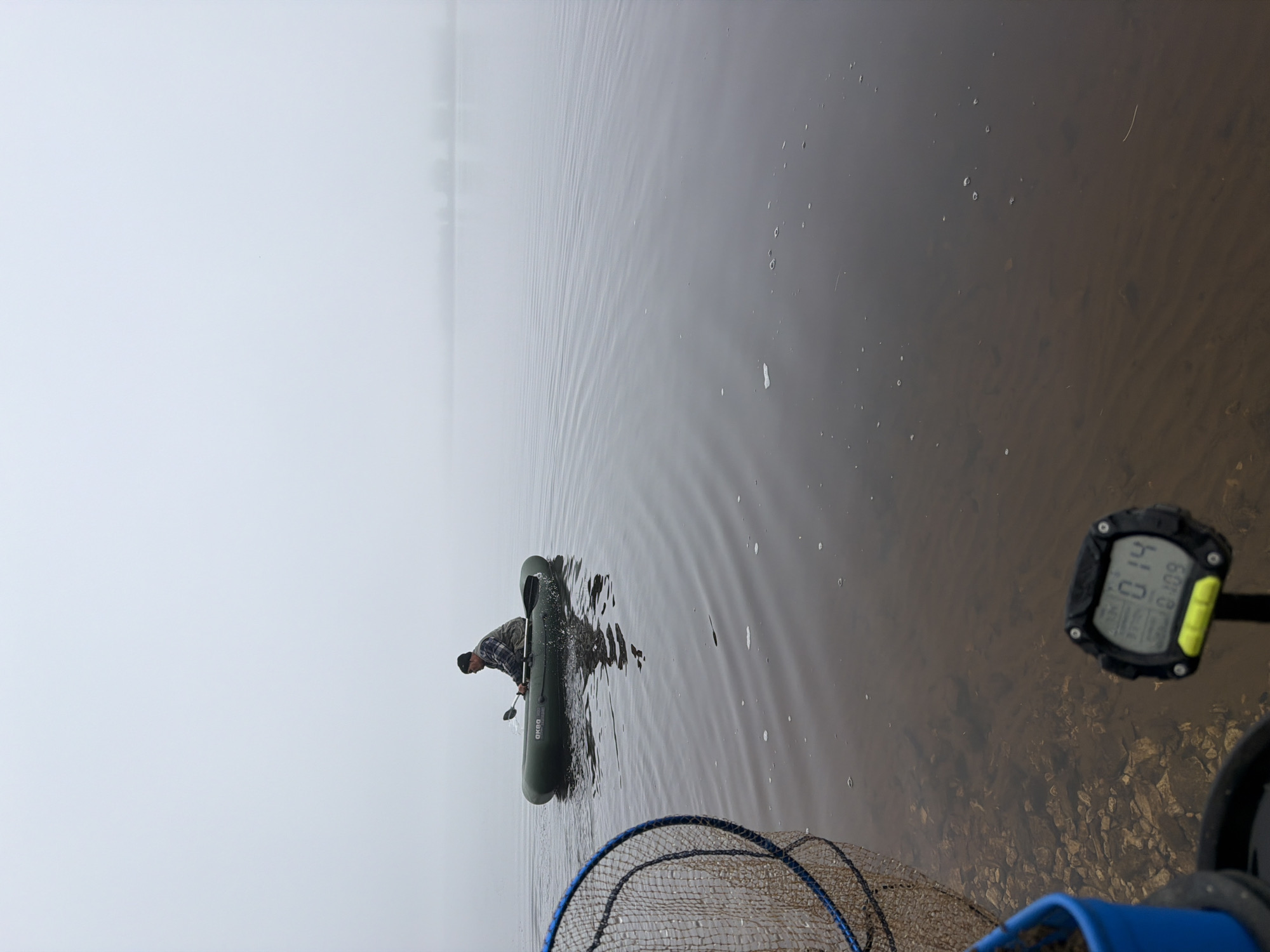 An outdoor scene captured on a foggy or misty day depicts a person in an inflatable boat on a calm body of water, likely a lake or river, in Chaikovsky, Russia. The image is rotated counter-clockwise.
In the corrected view, the foreground (bottom left) shows the edge of a blue-rimmed fishing net extending into the water. To the right, a digital device, possibly a fishfinder or a watch, is mounted, displaying the numbers "0.14" and "8:09", which could indicate time and a numeric reading like depth or speed. The water in this area is clear enough to reveal a rocky or pebbled bottom.
In the midground, a lone individual wearing a plaid shirt and a dark cap is positioned inside an olive green inflatable boat. The boat has the brand "BADGER" visible on its side. The person appears to be holding a paddle or a long pole, possibly maneuvering the boat or preparing to fish. Gentle ripples spread across the water surface around the boat.
The background is enveloped in thick fog or mist, obscuring the distant shoreline or any other landscape features, creating a serene and isolated atmosphere. The overall lighting is diffused and soft, consistent with an overcast and misty day. The calm water reflects the hazy sky, adding to the tranquil mood of the scene.