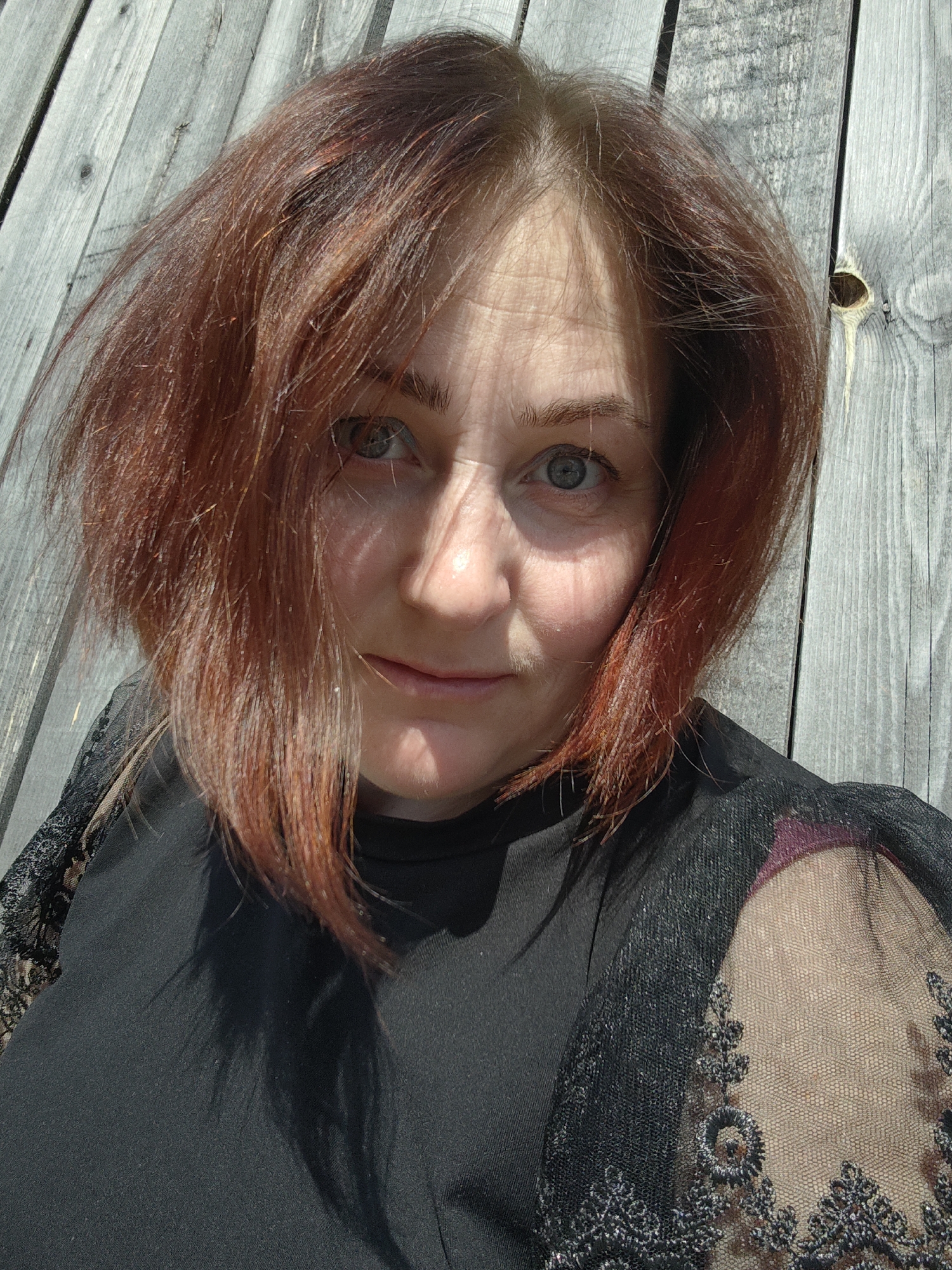 A middle-aged woman with auburn hair stands outside, facing the camera. She is wearing a black top with lace sleeves. The background consists of weathered, gray wooden planks. The lighting is bright, casting shadows across the woman's face. The image was taken outdoors, possibly in Фоки, Russia.