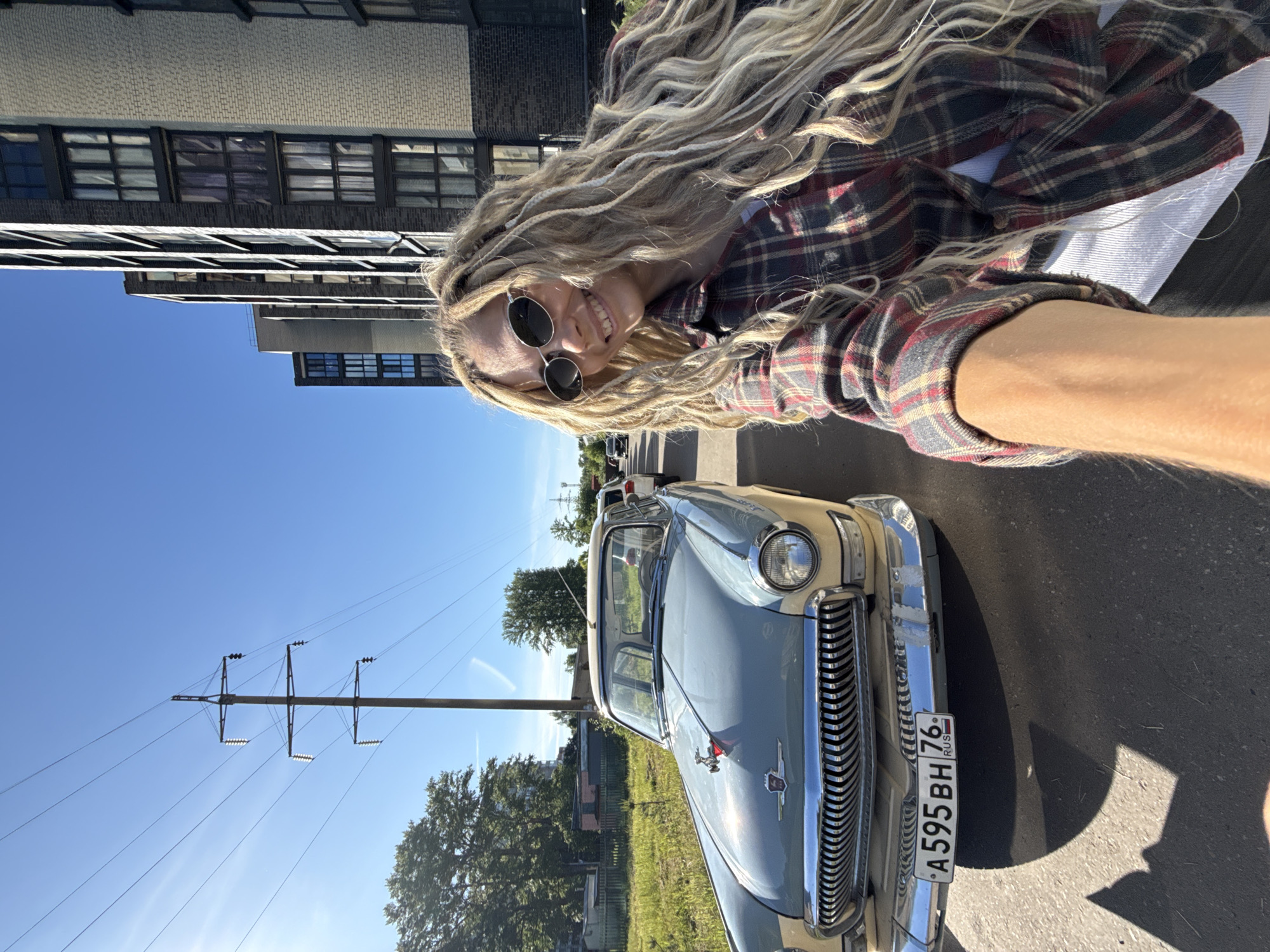 This bright, sunny image is a selfie taken by a smiling young woman with long, wavy blonde hair, some of which appears to be braided or dreaded. She wears round dark sunglasses and a red, black, and white plaid shirt over a white top. Her right arm, holding the camera, is visible in the lower right foreground.
Behind the woman, prominently featured, is a vintage light blue and beige car, identified as a GAZ-21 Volga by its distinctive front grille, chrome bumper, and hood ornament. A Russian license plate, "A595BH 76 RUS," is affixed to the front of the vehicle. The car is parked on asphalt pavement, which shows strong shadows, indicating bright daylight.
The background features a clear, deep blue sky. Several buildings are visible, including a light-colored brick structure and darker, multi-storied buildings with numerous windows, suggesting an urban or industrial environment. Green trees are scattered throughout the scene, and a tall electricity pylon with multiple power lines stands out against the sky. The time of day is clearly daytime, with strong sunlight. The woman's happy expression suggests she is enjoying the moment. The setting is Шебунино, Russia.
