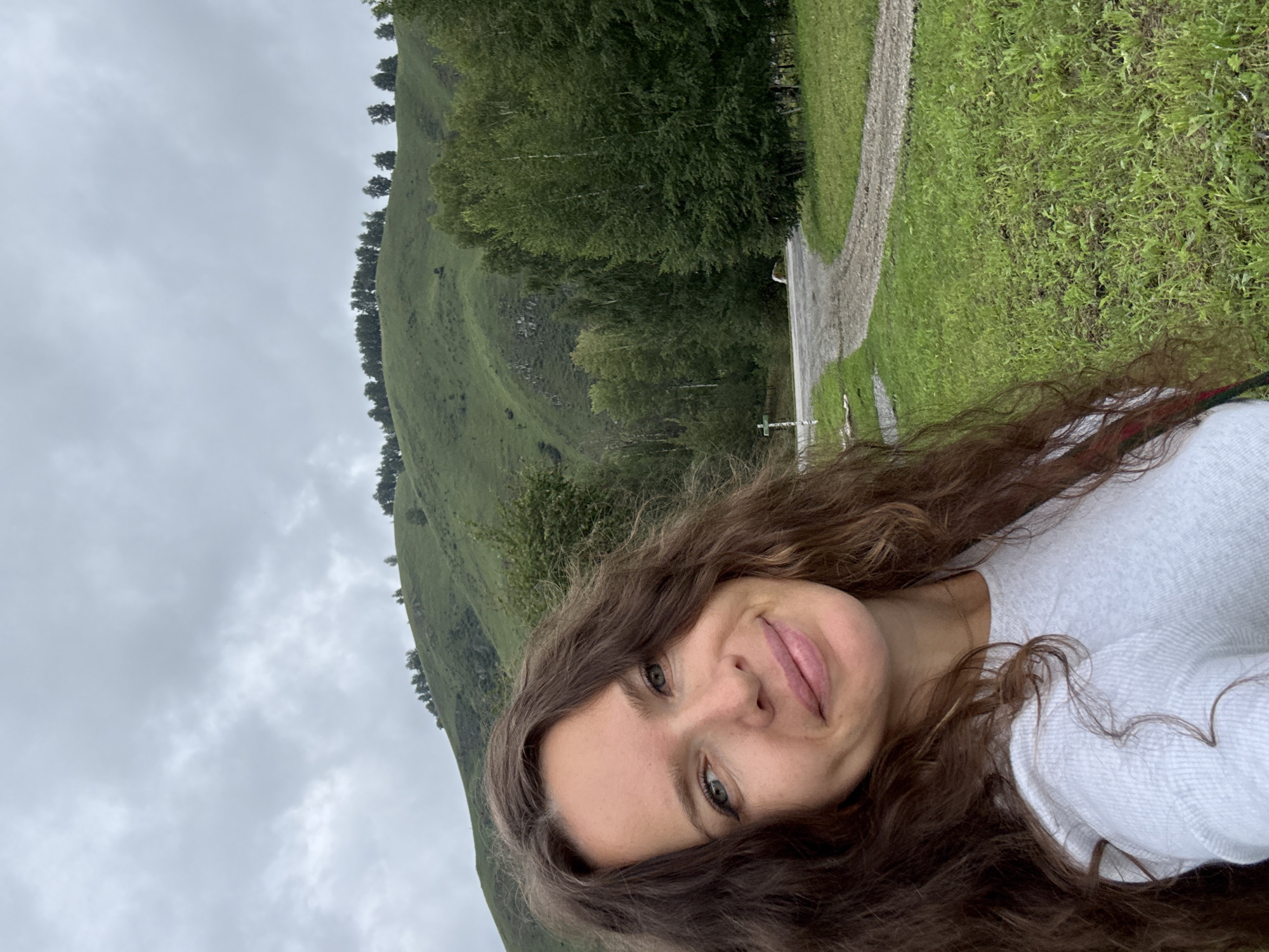 A woman with long, wavy brown hair is taking a selfie outdoors. She is wearing a light-colored, long-sleeved shirt. Her face is turned towards the camera, and she is smiling. Behind her, a green, treed hillside rises under a cloudy sky. A gravel road curves through the grassy foreground and into the treeline. The scene appears to be in a rural or natural setting.