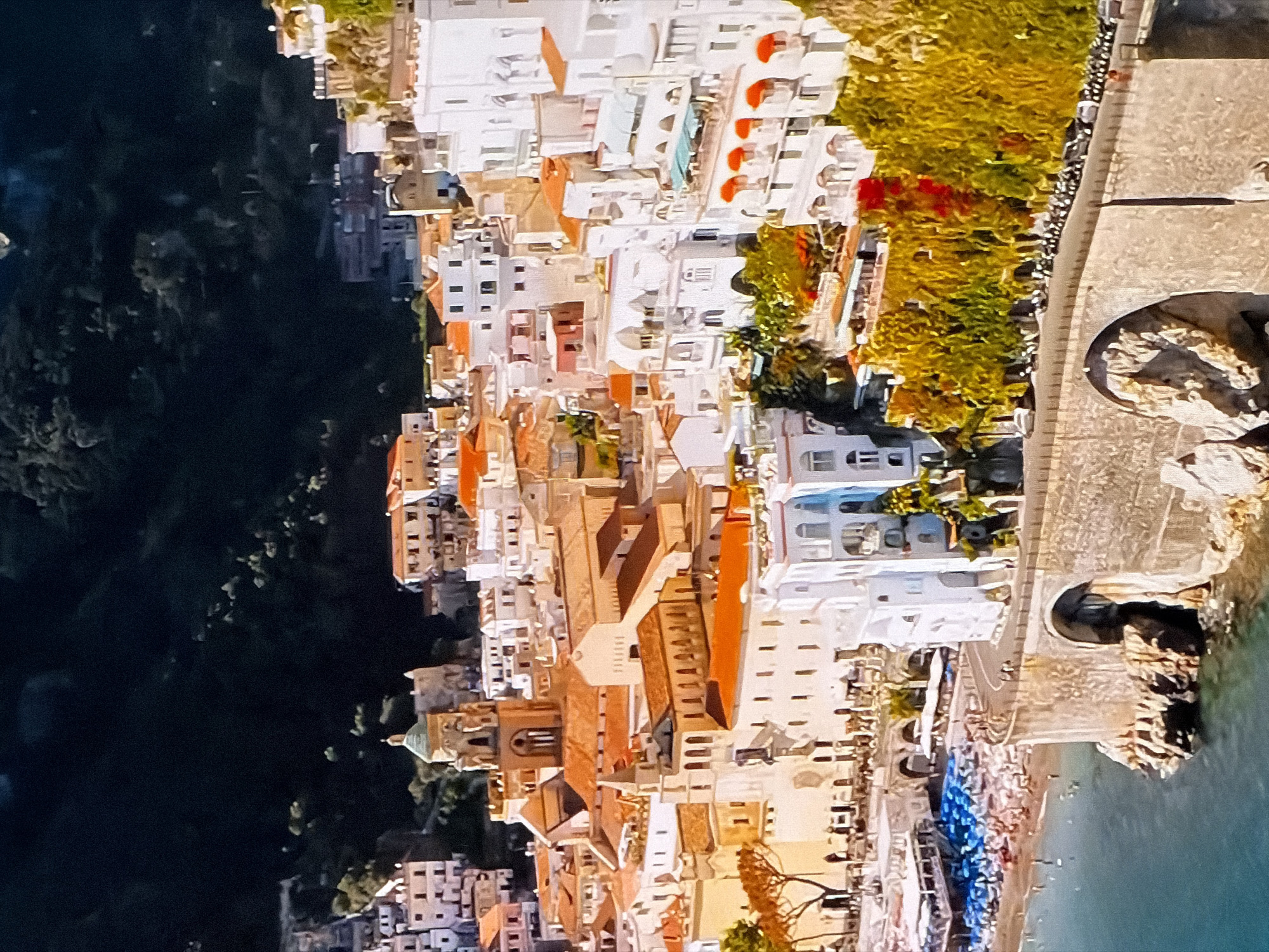 The image shows a scenic coastal town with white buildings, orange tiled roofs, and lush greenery. In the foreground, a stone bridge with arches spans over the water, which laps against rocky cliffs. Beyond the bridge, a beach is dotted with blue umbrellas and people. Further inland, buildings climb the hillsides, with a prominent church tower featuring a green dome. The dark, forested mountainside forms a dramatic backdrop to the vibrant town. This scene is of Amalfi, Italy.
