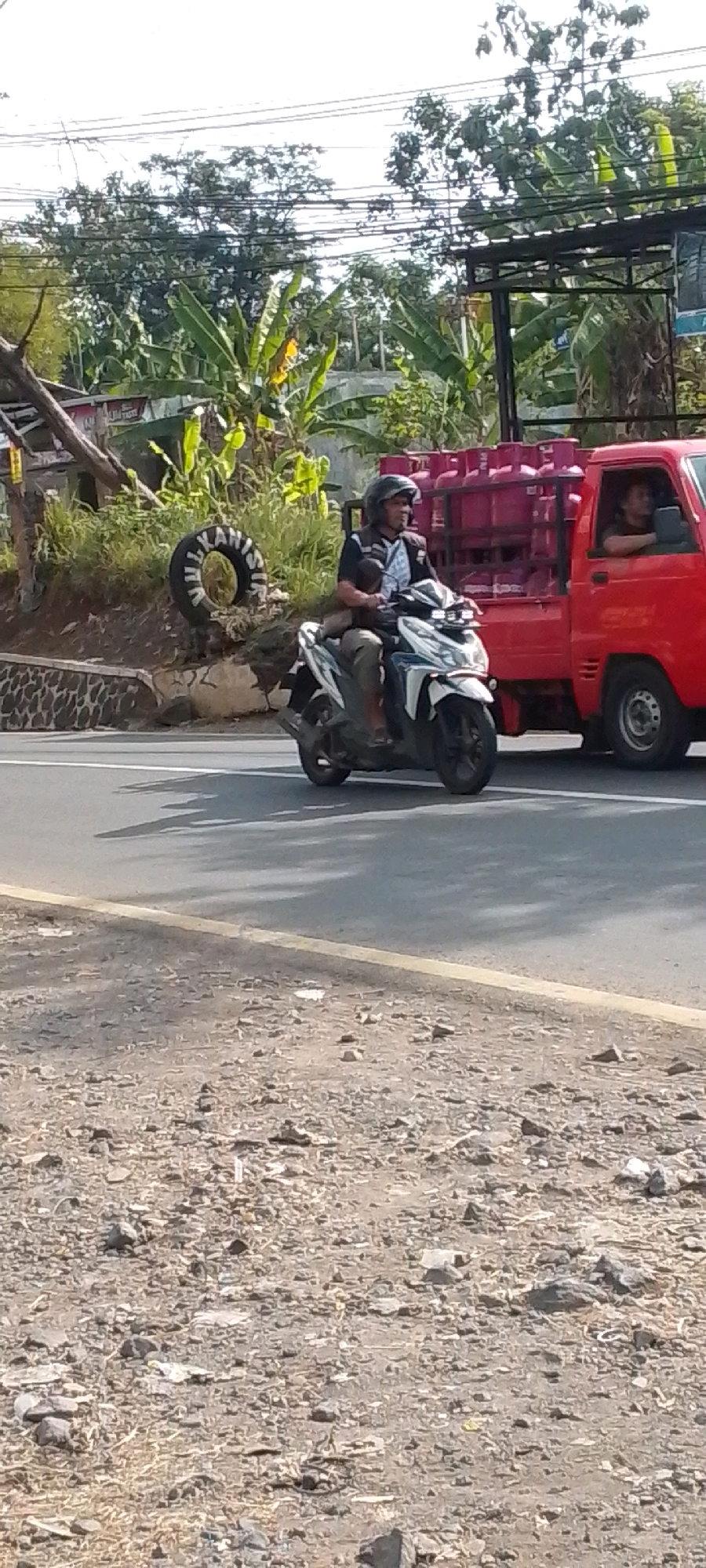 A man rides a white scooter on a paved road in Cilaku, Indonesia. He wears a black helmet and a dark shirt over a white and black patterned garment. He is positioned in the foreground, to the left of a red flatbed truck. The truck is loaded with several large, pink propane tanks. A second person, possibly the driver, is visible in the truck's cab. To the left of the road, there is a grassy embankment with a tire propped up against it. Behind the embankment are trees, including banana trees, and power lines. The scene appears to be set during the daytime, under a bright sky. The road surface is asphalt, with a yellow line marking the lane. The embankment on the left is covered in gravel and small stones.