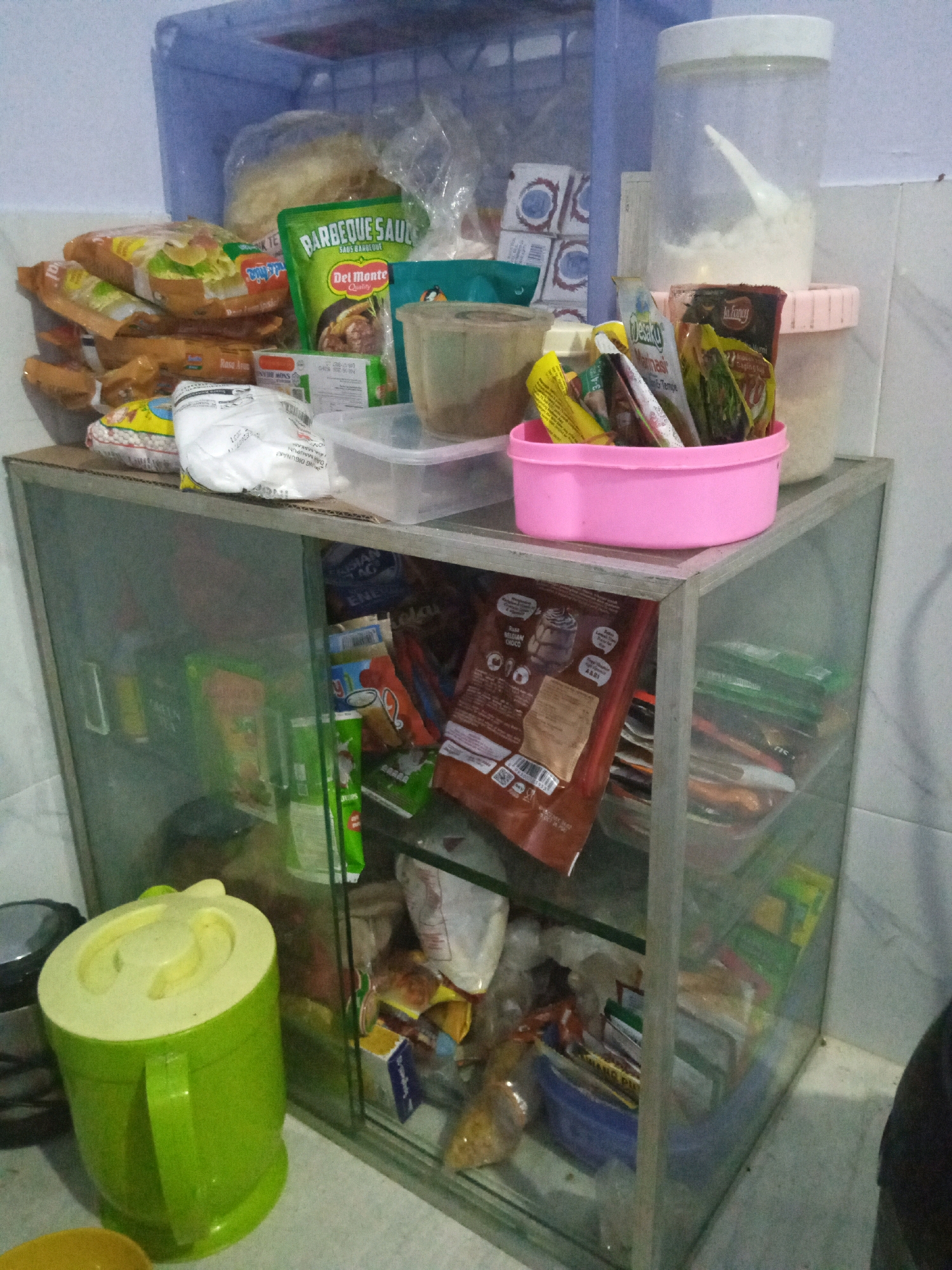 The image shows a shelf unit filled with food items and containers. The shelf unit is made of glass and metal. On the top shelf, there are various packaged food items, including what appears to be instant noodles, a bottle of barbecue sauce, and packets of spices. There is also a clear plastic container with a white lid, filled with a white powdery substance. To the right, a pink plastic container holds several small packets of spices and seasonings.
On the shelves below, more food items are visible, including packets of crackers, snacks, and other packaged goods. A large green plastic container with a lid is placed on the surface to the left of the shelf unit. The environment appears to be a kitchen or pantry area, with tiled walls in the background. The lighting is somewhat dim. The image does not contain any people or any indication of activity. It is a static scene of stored food items. The location is identified as Sumbergirang, Indonesia.