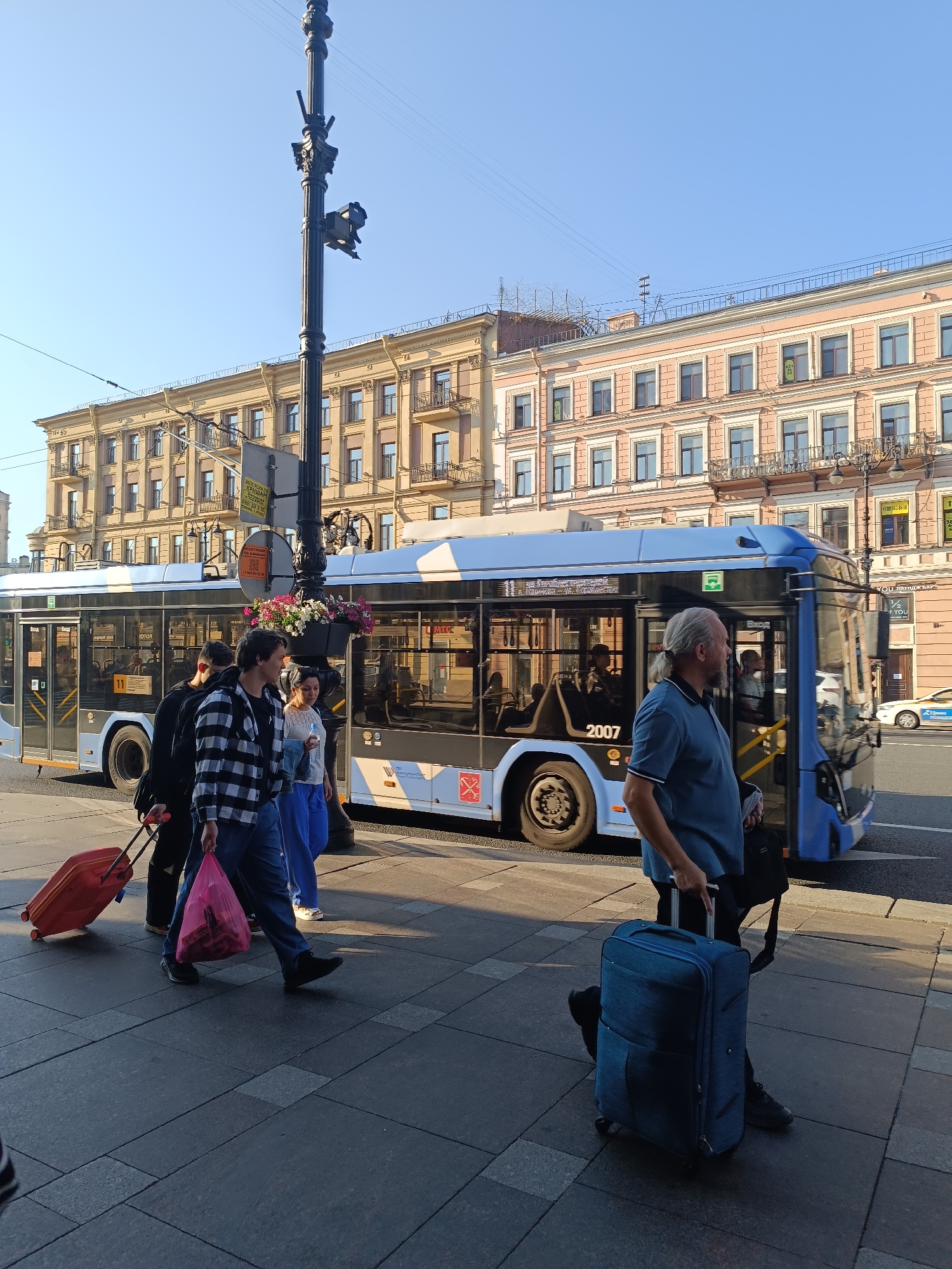 The image depicts a bright, sunny day on a city street in Saint Petersburg, Russia. In the foreground, a group of people are walking on a pedestrian sidewalk. To the left, a person is pulling a red suitcase. Next to them, another person in a black and white checkered shirt is carrying a pink plastic bag. Behind them, a third person is partially visible. Further to the right, a man with long grey hair tied back in a ponytail is standing next to a large blue bus, holding a black bag in his left hand and a blue suitcase with wheels in his right.
The bus, identified by the number 2007 on its side, is a modern city bus with tinted windows. It has "МЕТРО" (METRO) written on it in Russian. Several people are visible through the windows of the bus. A decorative planter with flowers is attached to a lamppost in front of the bus. The lamppost itself is ornate and dark.
In the background, tall, multi-story buildings with classical architecture line the street. These buildings are painted in light colors, possibly yellow and pink, and feature numerous windows and balconies. Wires are strung overhead, indicating the presence of the city's electrical infrastructure, possibly for trams or trolleybuses. The sky is clear and blue. The scene appears to be a typical urban environment in Saint Petersburg during the daytime.