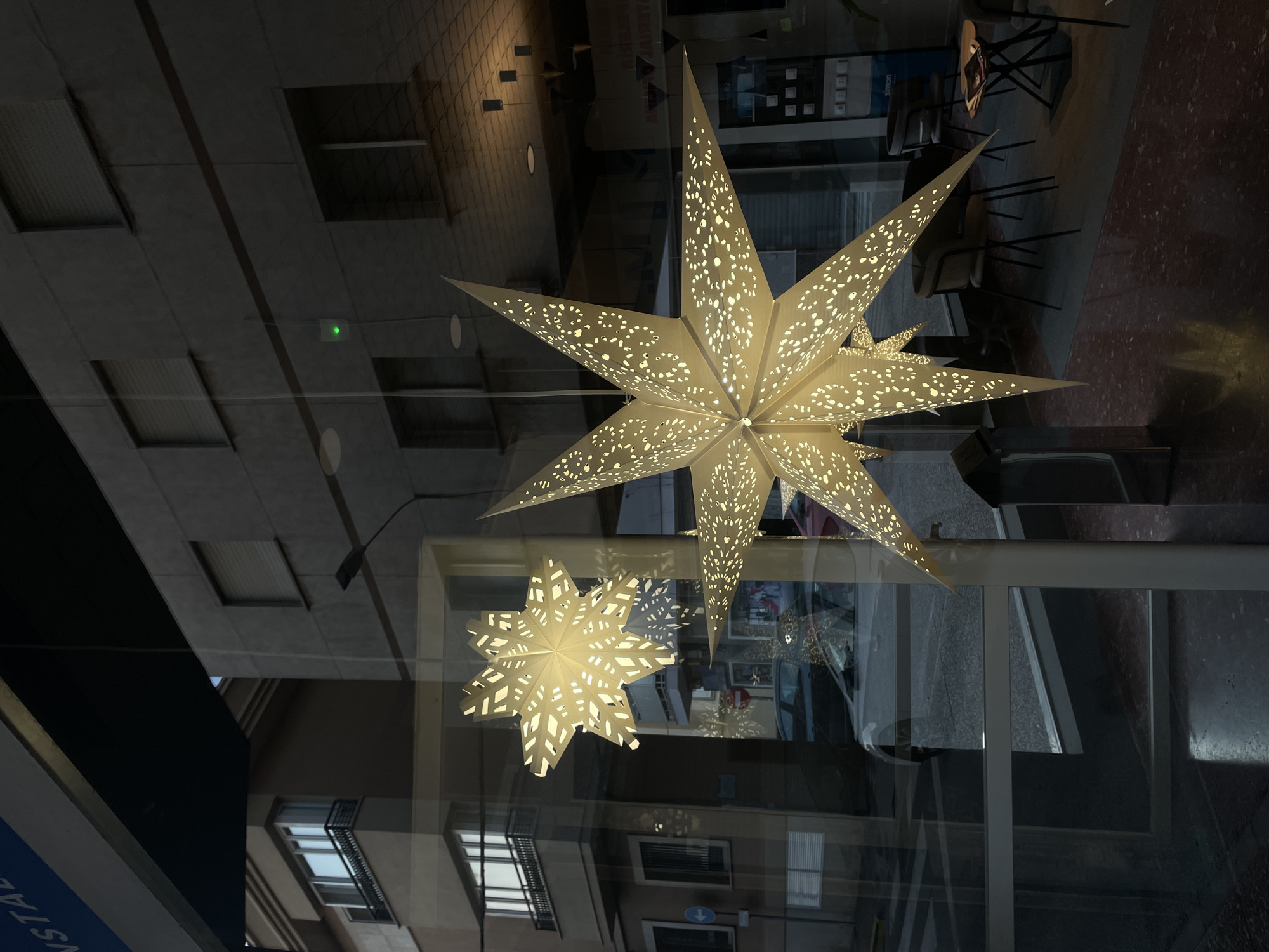 The image is a medium-angle shot of a storefront window on a street in La Línea de la Concepción, Spain. Two illuminated, paper star-shaped decorations are hanging in the window, casting intricate patterns of light onto the interior and reflecting the street scene outside. The larger star is in the foreground, partially obscuring the smaller star behind it.
Through the window, the exterior shows a gray, modern building with multiple stories and regularly spaced windows, some with closed blinds. A street with a crosswalk is visible, along with parked cars. The interior of the store is dimly lit, with a few chairs and tables visible, suggesting it might be a cafe or retail space. The floor inside has a speckled red and black pattern. A blue awning with white text, partially visible at the top of the image, indicates the establishment's signage. The overall impression is of a street-level commercial space, possibly decorated for a holiday.
