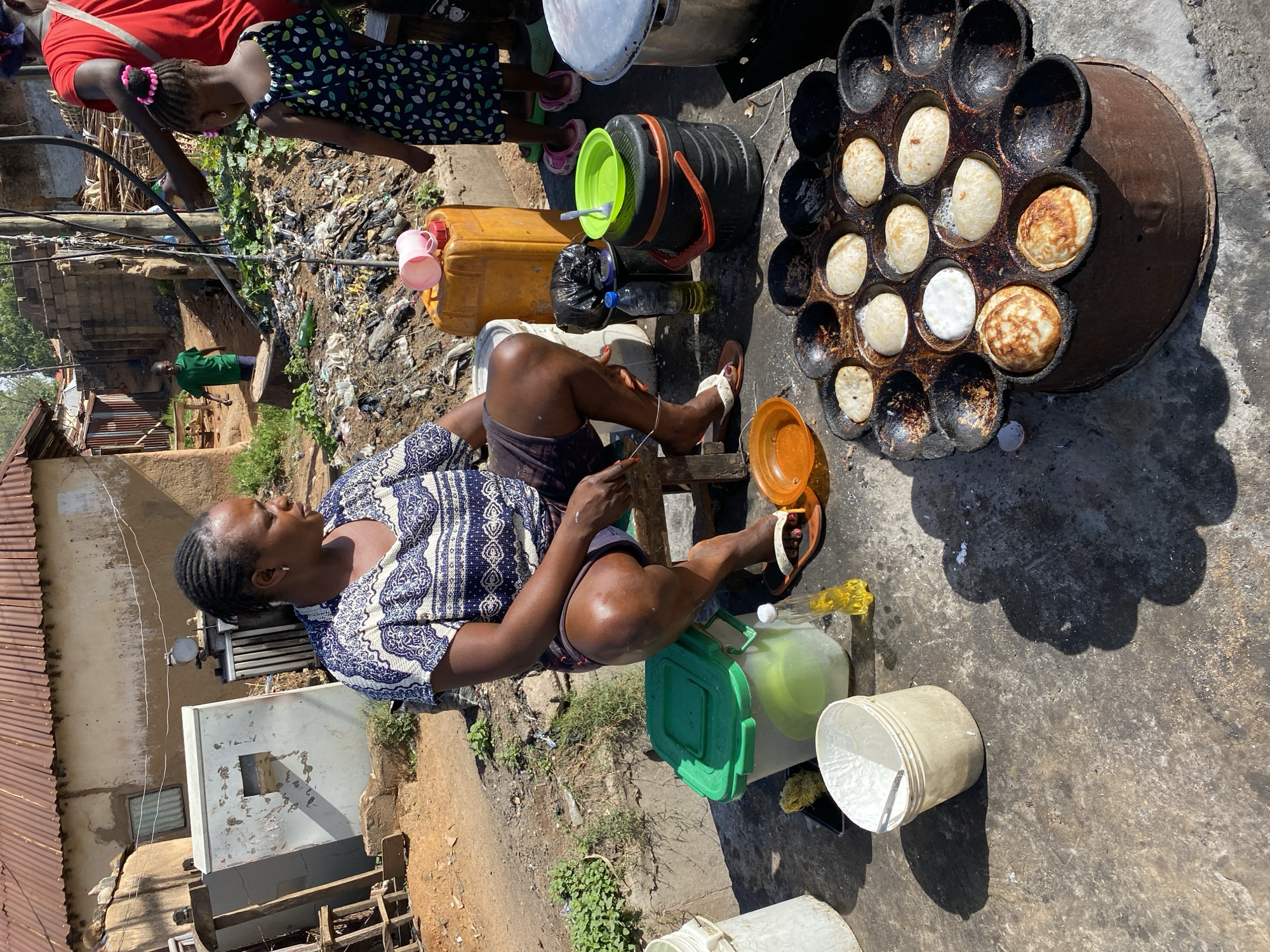 An eye-level, outdoor shot in Bukuru, Nigeria, features a woman sitting on a small, wooden stool beside a large, round, multi-compartment cooking apparatus containing small, pancake-like items. The woman, attired in a blue and white patterned shirt and dark shorts, is engaged in an unidentified activity with an orange container, her left leg crossed over her right. The cooking apparatus sits atop a rusty metal drum.
Adjacent to the woman are several containers, including a green-lidded plastic container and a yellow jerrycan. Behind her are several items, including a black cylindrical container with a green lid and a black trash bag. To the left is a building, a cluttered environment, and a child in a patterned dress. The ground appears to be a worn, concrete surface. Buildings and various objects scattered across the scene suggest an urban or semi-urban setting.
