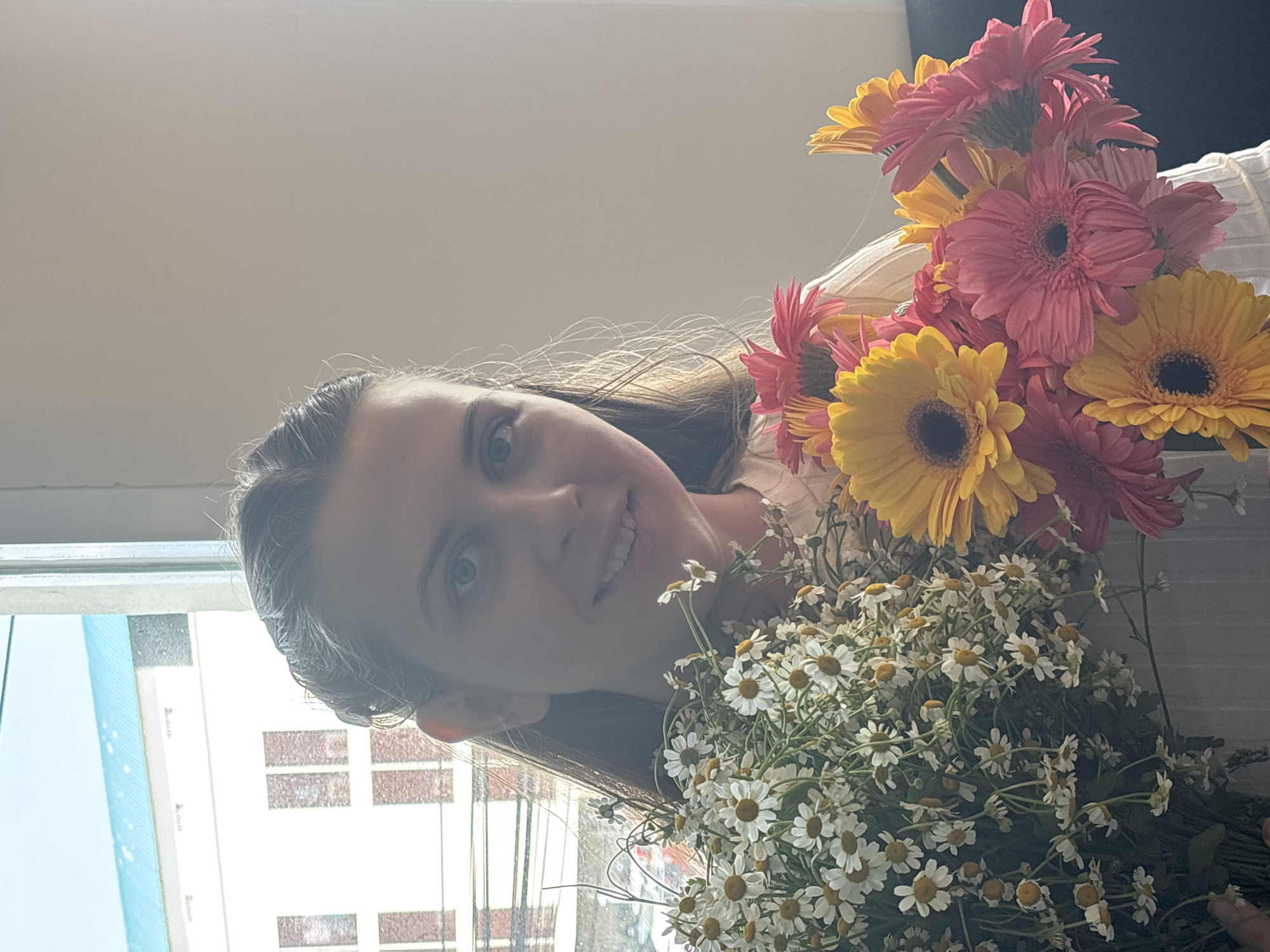A woman is holding two bouquets of flowers in front of her. One bouquet consists of white daisies with yellow centers, while the other is composed of pink and yellow gerbera daisies. The woman is smiling at the camera. The background features a window overlooking a building in Phuket City Municipality, Thailand.