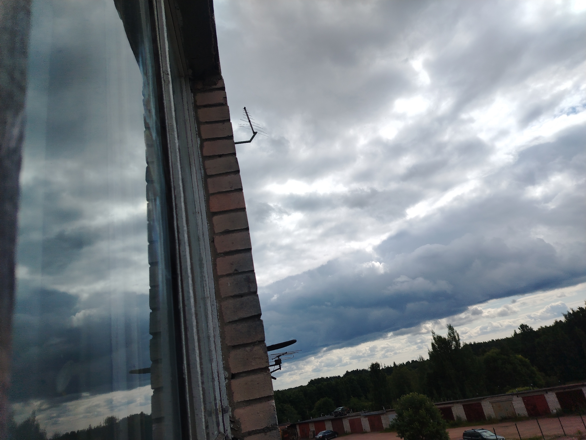 The image shows a view from a window looking outwards towards a cloudy sky and a line of garages. The window frame is made of brick, and the glass reflects the sky. An antenna is visible mounted on the brick wall. Below the sky, a dense line of trees is present, with a row of garages partially visible behind them. Two cars are parked in front of the garages. The overall impression is of an overcast day in an urban or suburban setting. The question states the location is Vyborg, Russia, but there are no visual cues within the image to confirm or deny this.