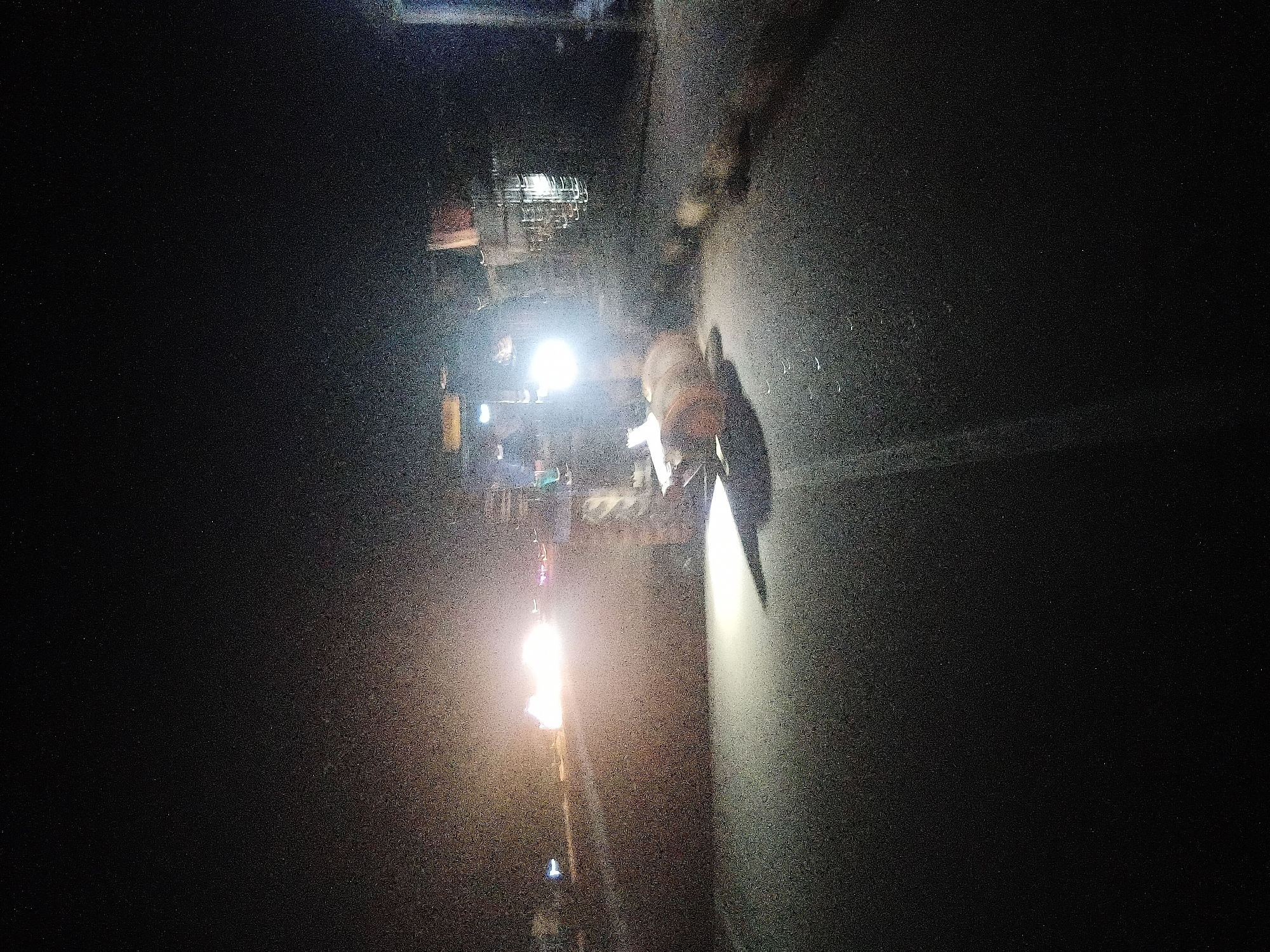 A tractor is seen on a road at night, with its headlights illuminating the scene. The tractor appears to be pulling a large, round object, possibly a hay bale or a piece of equipment. The road is paved and has a white line marking the center. In the background, there are other vehicles and lights, suggesting an urban or semi-urban environment. The overall impression is one of nighttime agricultural or construction work taking place on a road in Jalingo, Nigeria.
