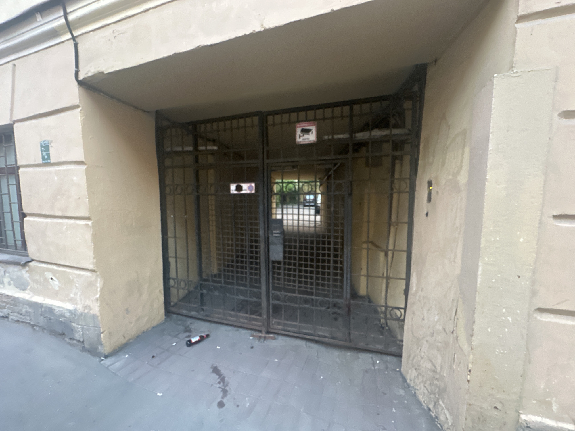 A street-level, medium shot depicts the entrance to an apartment building or courtyard in what appears to be Saint Petersburg, Russia. The entrance is framed by weathered, light-beige colored walls and an overhead archway. A black metal gate, secured with an unknown locking mechanism, blocks access to the interior. A sign indicating CCTV monitoring is fixed to the gate. Litter, including a small bottle, is visible on the gray tiled pavement in front of the gate. The environment appears urban and somewhat neglected.