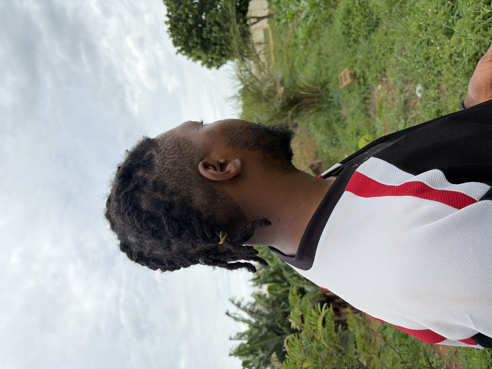 A close-up, profile view captures a man from the shoulders up, looking to his right. He has dreadlocks styled into a ponytail secured with a yellow band, and a short beard. He is wearing a black shirt with white and red accents. The background is a lush, green outdoor environment with various plants and trees, including banana trees and palm trees. The sky is overcast with clouds. The scene appears to be in Rafin Bauna, Nigeria.