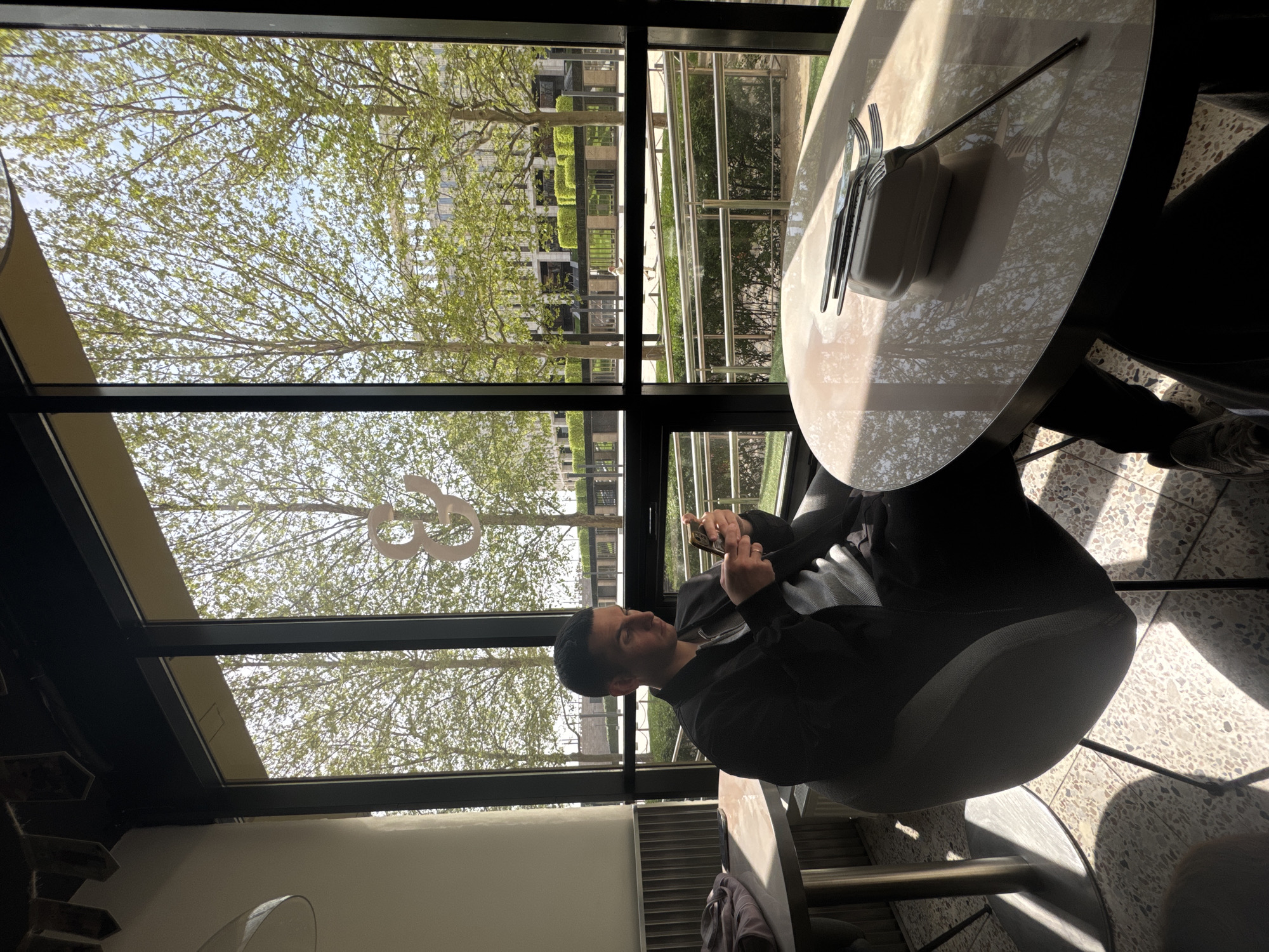 A man is seated at a round table next to a large window, looking at his phone. He is dressed in dark clothing, possibly a black jacket or shirt, and has short dark hair. A ring is visible on his left hand as he holds the device with both hands. The table is light-colored, possibly marble or a similar polished surface, and holds a container with several forks. Strong reflections of the tree canopy and window frames are visible on the table's surface.
The setting appears to be a cafe or restaurant with extensive glass windows and a glass ceiling or skylight, allowing abundant natural light to flood the space. Outside the window, a vibrant green canopy of trees, likely deciduous, indicates springtime or summer. Through the trees, modern-looking buildings are partially visible in the background, suggesting an urban or park-like environment within a city. The floor is tiled with a speckled pattern, and distinct shadows from the window frames and furniture are cast across it, indicating a bright, sunny day, likely morning or late afternoon given the angle of the light. On one of the window panes, a stylized "G3" or similar character is visible. The atmosphere is bright and airy.