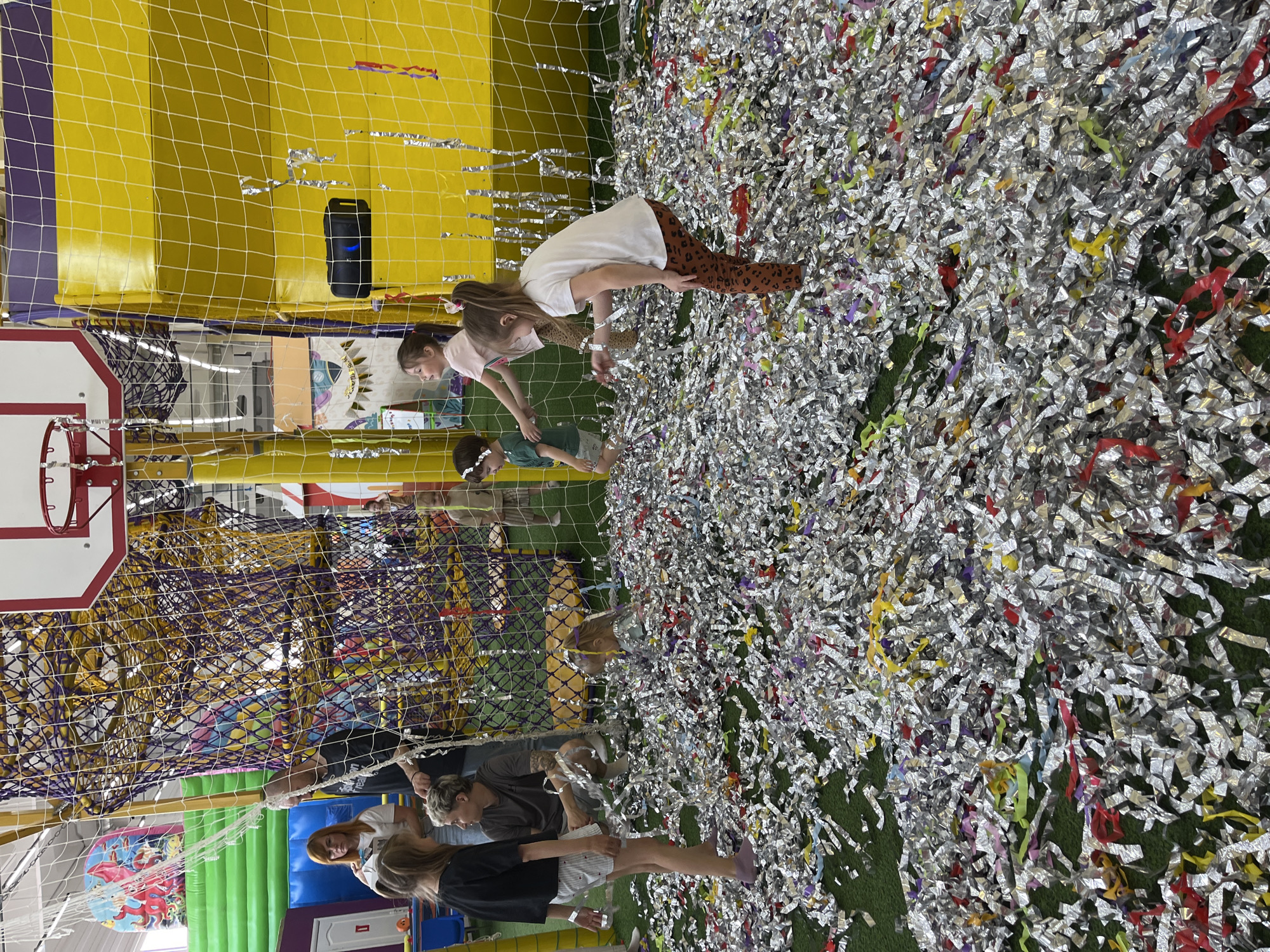 The image depicts an indoor playground, likely in Chaikovsky, Russia. Three children are playing in a large pit filled with silver, yellow, red, and purple confetti strips on a green base. A net separates the confetti pit from another play area with yellow and purple padding. A basketball hoop is visible in the corner. Behind the net, adults are standing.