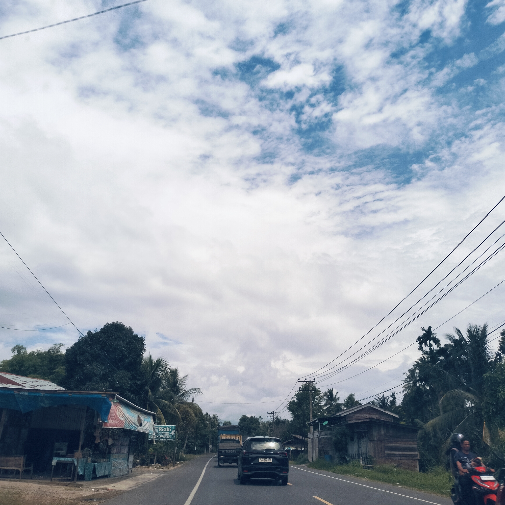 The image captures a rural or semi-rural road scene in Pandrahkandeh, Indonesia, under an overcast sky with patches of blue. The upper half of the image is dominated by the sky, filled with a mix of white and light blue clouds.
A two-lane asphalt road with white and yellow road markings stretches through the center of the frame, leading away from the viewer. In the mid-ground, a black SUV is visible from the rear, its license plate reading "BL 1330 AAF". Ahead of it, a light-colored truck, possibly a Mitsubishi, can be seen with "MITSUBISHI" printed on its tailgate. Both vehicles are moving away from the viewer. On the right side of the road, a red motorcycle with two occupants, one wearing a helmet, is driving towards the viewer.
The sides of the road are lined with lush tropical vegetation, including numerous tall palm trees and dense green foliage. On the left side, there's a simple, open-front roadside stall or shop with a blue tarp roof and a faded red, white, and blue banner. A smaller blue sign below the banner clearly displays the word "Rizki" and features an image of a truck. Further along the left, another building is partially obscured by trees. On the right, a wooden house with a red roof is visible amidst the trees. Multiple black power lines are strung across the sky, connecting utility poles on both sides of the road. The scene suggests a typical day with ongoing transit and local commerce in a tropical, rural setting.