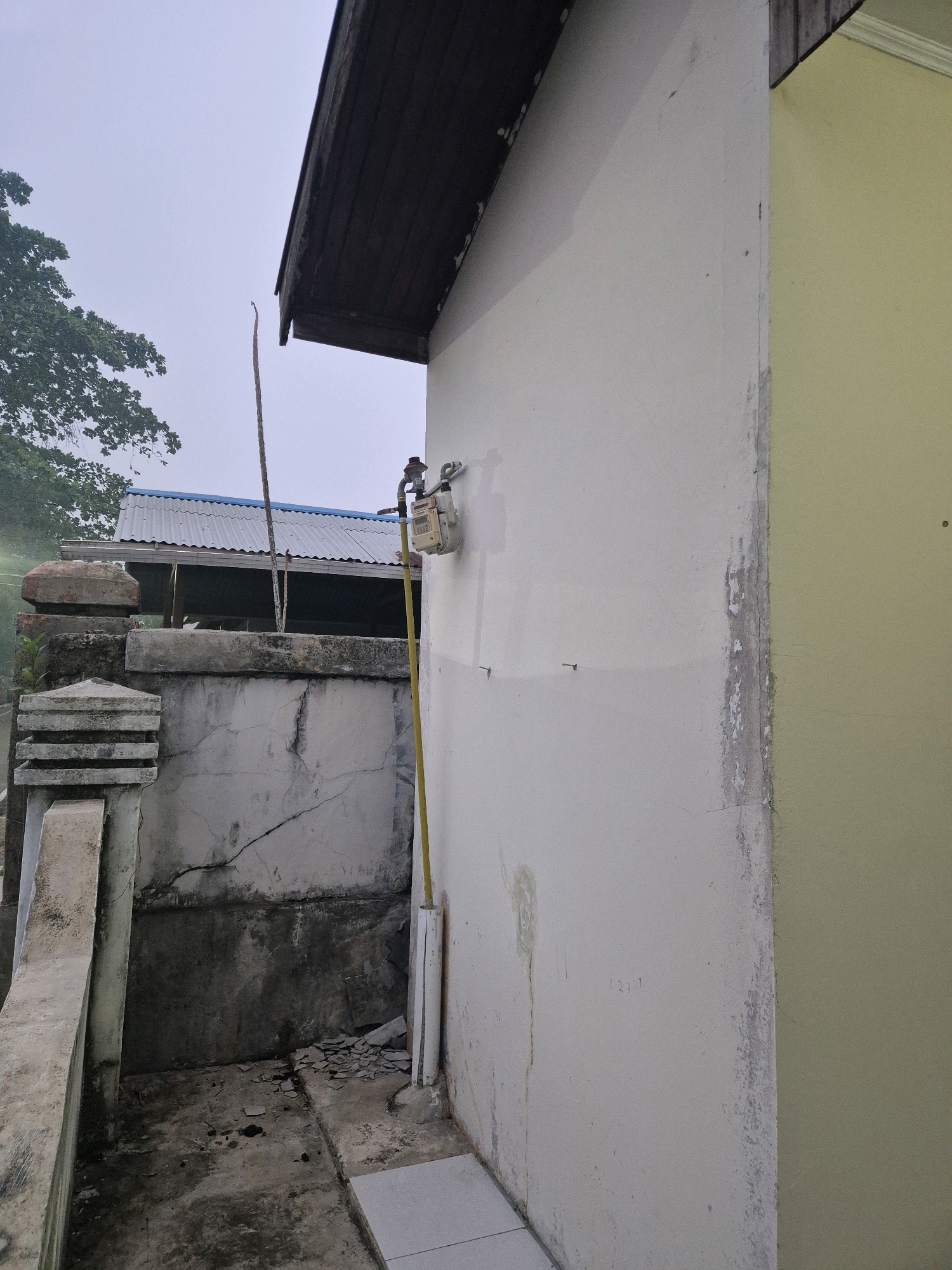 The image depicts an exterior view of a residential building in Bontang, Indonesia, under an overcast sky. The foreground features a light-colored wall with a gas meter mounted on it, connected to a yellow pipe running vertically down the wall. To the left, a cracked concrete wall and structure are visible, along with debris on the ground. In the background, a corrugated metal roof structure is partially obscured by a tree. The scene appears to be an outdoor area, possibly a backyard or side yard, with signs of wear and tear on the concrete elements.
