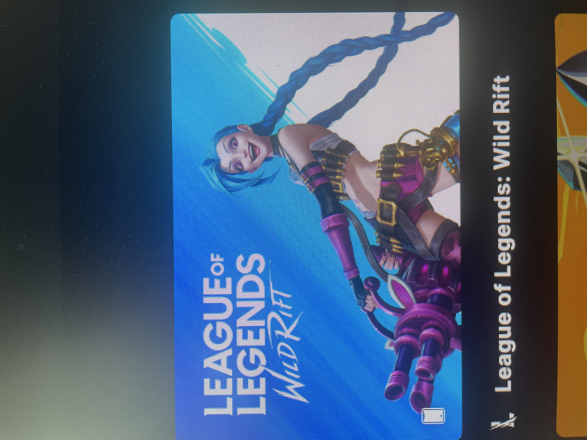 The image shows a screenshot of a game's title card, featuring the "League of Legends: Wild Rift" logo and artwork of the character Jinx. The logo is in white text against a blue background. Jinx is depicted holding a large, pink weapon, with her signature blue braids and a mischievous grin. Below the title card, the full title "League of Legends: Wild Rift" is displayed in white text on a black background. The image is a photograph of a screen, indicated by the pixelation and slight glare. The location is not identifiable in the image.