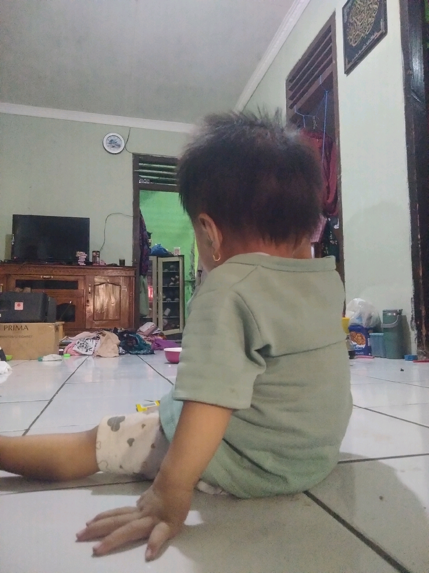 The image depicts an interior scene, likely a living room, in Suka Sari, Indonesia. A young child, viewed from behind, is sitting on a tiled floor. The child is wearing a light green shirt and light-colored shorts with a pattern of grey hearts. Their left arm is extended on the floor, with their hand open.
The environment includes a wooden entertainment center with a dark television on top. To the left of the entertainment center is a large brown box with "PRIMA" printed on it. Various items of clothing are scattered on the floor in front of the entertainment center. Further back, a wooden cabinet with glass doors, possibly a shoe rack, is visible, containing shoes. A doorway or opening leads to another room with green walls.
On the wall above the entertainment center, a round clock is visible. To the right of the doorway, another framed artwork hangs on the wall. In the background, near the doorway, a blue bag and a green bucket are present. The lighting suggests it is daytime, possibly with natural light filtering in from the adjacent room. The overall impression is of a casual, lived-in domestic space.