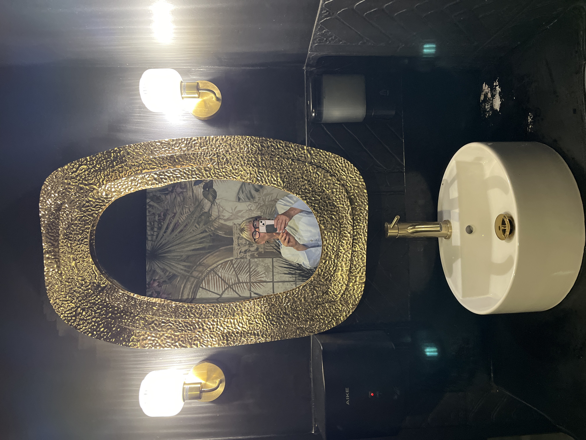 The image depicts the interior of a bathroom. The walls are dark, possibly black, with some areas showing a textured pattern. A gold-colored, ornate oval mirror hangs on the wall, reflecting a person taking a photograph with a mobile phone. Two light fixtures with round bulbs are mounted on either side of the mirror. To the right of the mirror is a white, round sink with gold-colored fixtures. Below the mirror is an "AIKE" branded hand dryer, and above the sink is a soap dispenser. The image was taken in Saint-Jean-de-Védas, France.