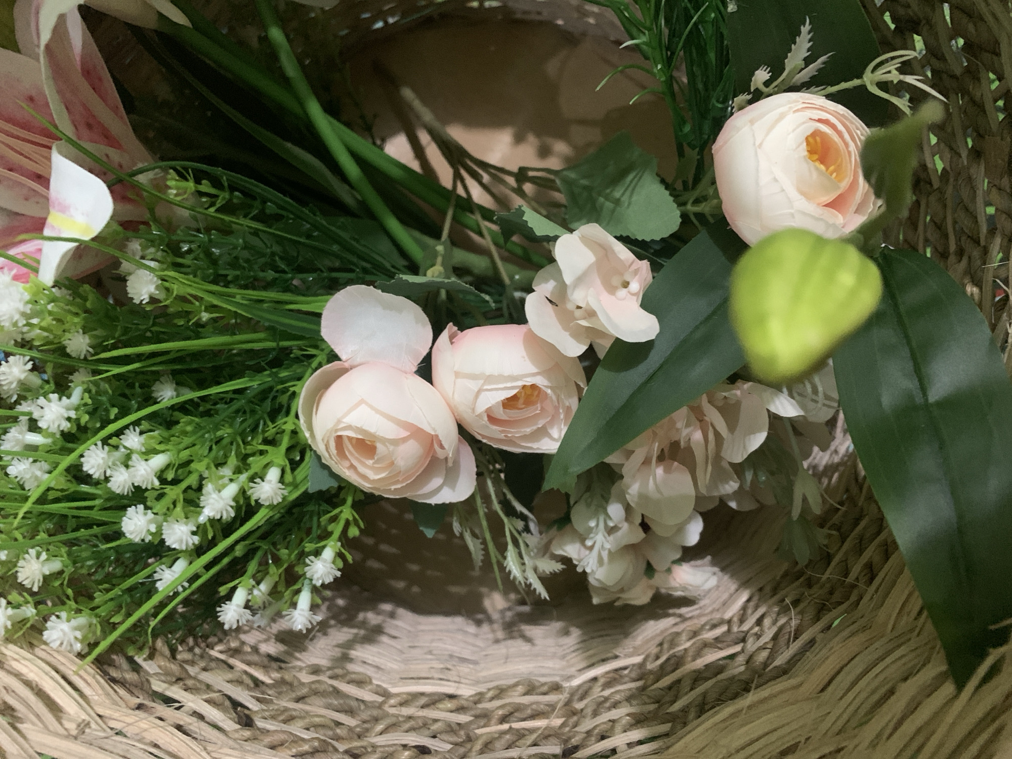 This is a close-up, eye-level shot of a floral arrangement in a woven basket. The arrangement features several light pink roses, smaller white flowers that resemble baby's breath, and green foliage. One prominent light pink rose is in the lower right portion of the frame, with its petals tightly furled. Another similar rose is above and to the left of it. A cluster of the white flowers is visible in the upper left. The basket appears to be made of a natural, fibrous material with a thick, braided texture. The background is a neutral, shadowy brown, suggesting an indoor setting. The lighting is soft, casting gentle shadows. The scene is static, with no visible action. The image is likely a still life composition. There are no people or animals present. The location in Kuala Pembuang, Indonesia, is not discernible from the visual information provided in the image.