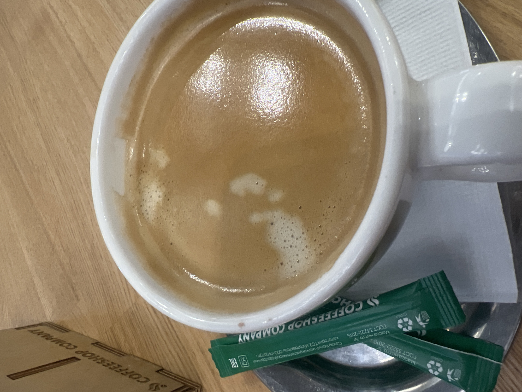A close-up, overhead shot shows a white mug filled with a frothy, brown coffee. The mug sits on a silver saucer, which is placed on a light brown wooden table. Two green sugar packets, labeled "COFFEESHOP COMPANY," are positioned on the left side of the saucer. In the background, a brown cardboard box with "COFFEESHOP COMPANY" printed on it is partially visible. The lighting creates highlights on the coffee and the mug's handle.