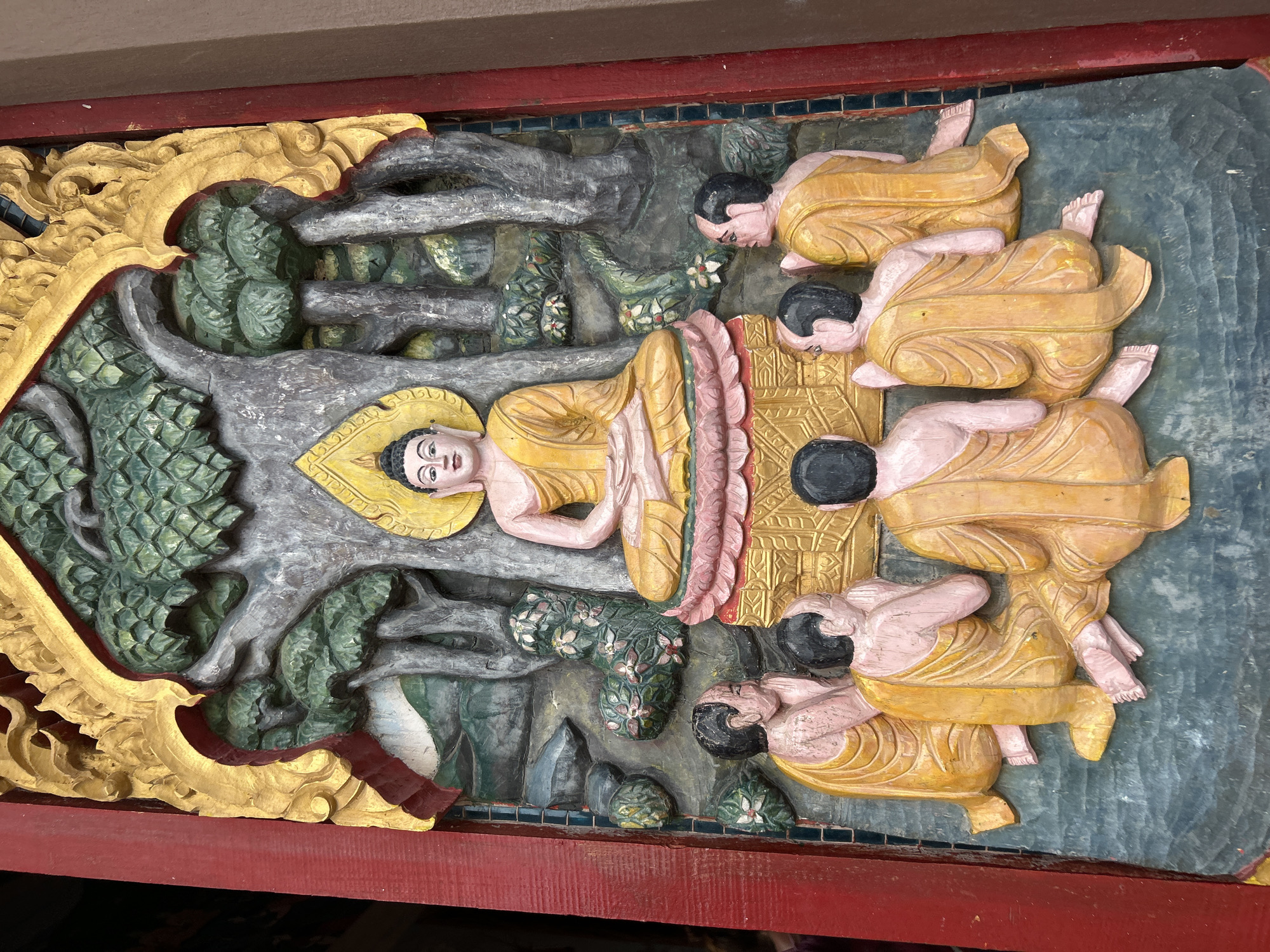 This is a detailed, low-angle, medium shot of a carved wooden relief depicting a Buddhist scene, likely located in a temple or shrine within Ko Samui District, Thailand. The artwork is vibrantly colored, with a prominent Buddha figure seated centrally under a stylized tree. The Buddha is depicted in meditation, wearing saffron robes and with a halo. Surrounding the Buddha are five monks, also in saffron robes, kneeling and appearing to be in prayer or deep contemplation. The background features a stylized forest with a body of water and scattered foliage. The relief is framed by a red wooden structure on the left and right, with ornate golden trim along the top edge. The overall impression is one of religious devotion and artistic craftsmanship.