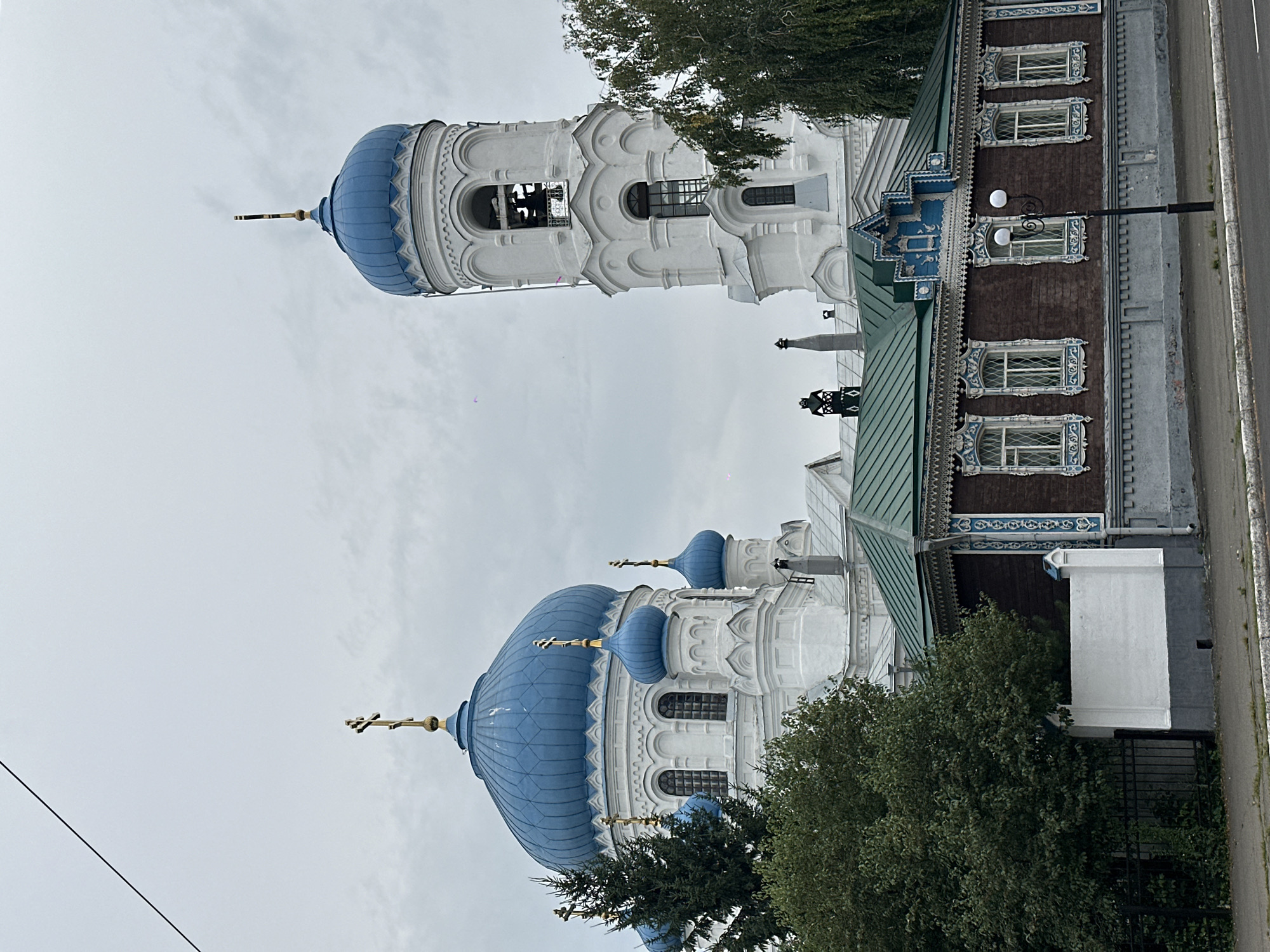 The image depicts an overcast day in Biysk, Russia, focusing on the majestic Uspensky Cathedral and a traditional wooden house. The cathedral, a prominent white structure with striking blue domes, dominates the background. Its main dome is large and bulbous, topped with a golden cross. Several smaller blue domes with crosses are also visible, adding to its architectural grandeur. To the right, a tall, white bell tower rises, featuring arched openings and a bell within. In the foreground, a low, dark brown wooden house stands out with its ornate white trim around the windows and roofline, showcasing intricate carvings. The roof of the house is a vibrant green. A streetlamp with two globes stands near the house. Green trees partially obscure the view of both the house and the cathedral. A dark metal fence and a white wall are visible to the left of the house, bordering a sidewalk. The sky is uniformly gray, suggesting a cloudy or potentially rainy day. No people are visible in the image.